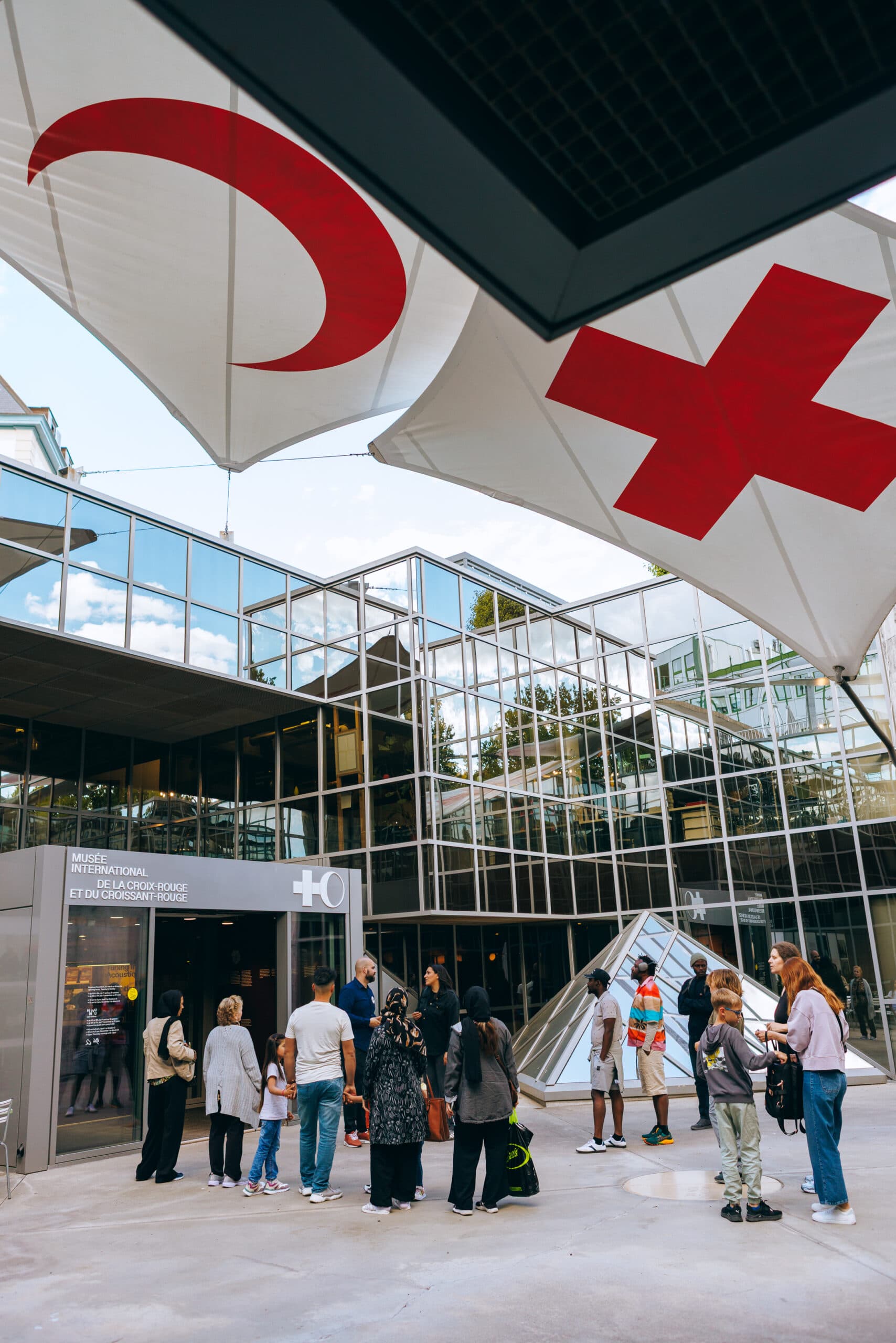 Glass façade of a modern building with two large flags of the Red cross and Red Crescent emblems hanging above the entrance, with people walking inside.