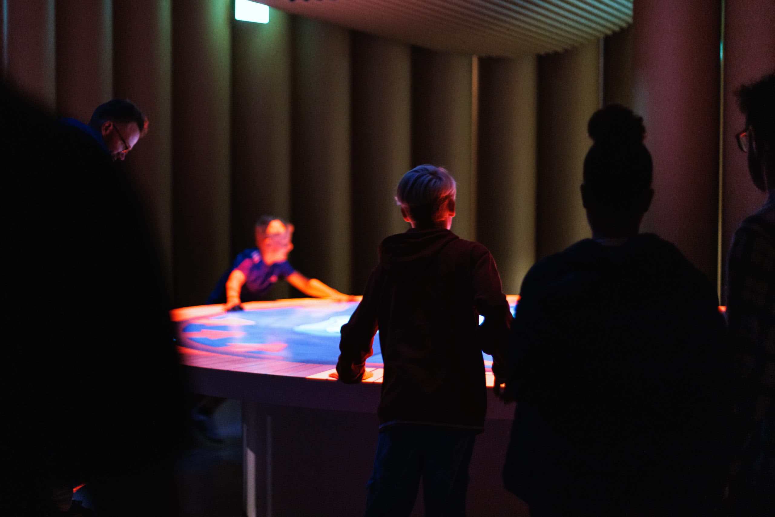 Several visitors are in a dark room, gathered around a light table onto which an interactive game is being projected.