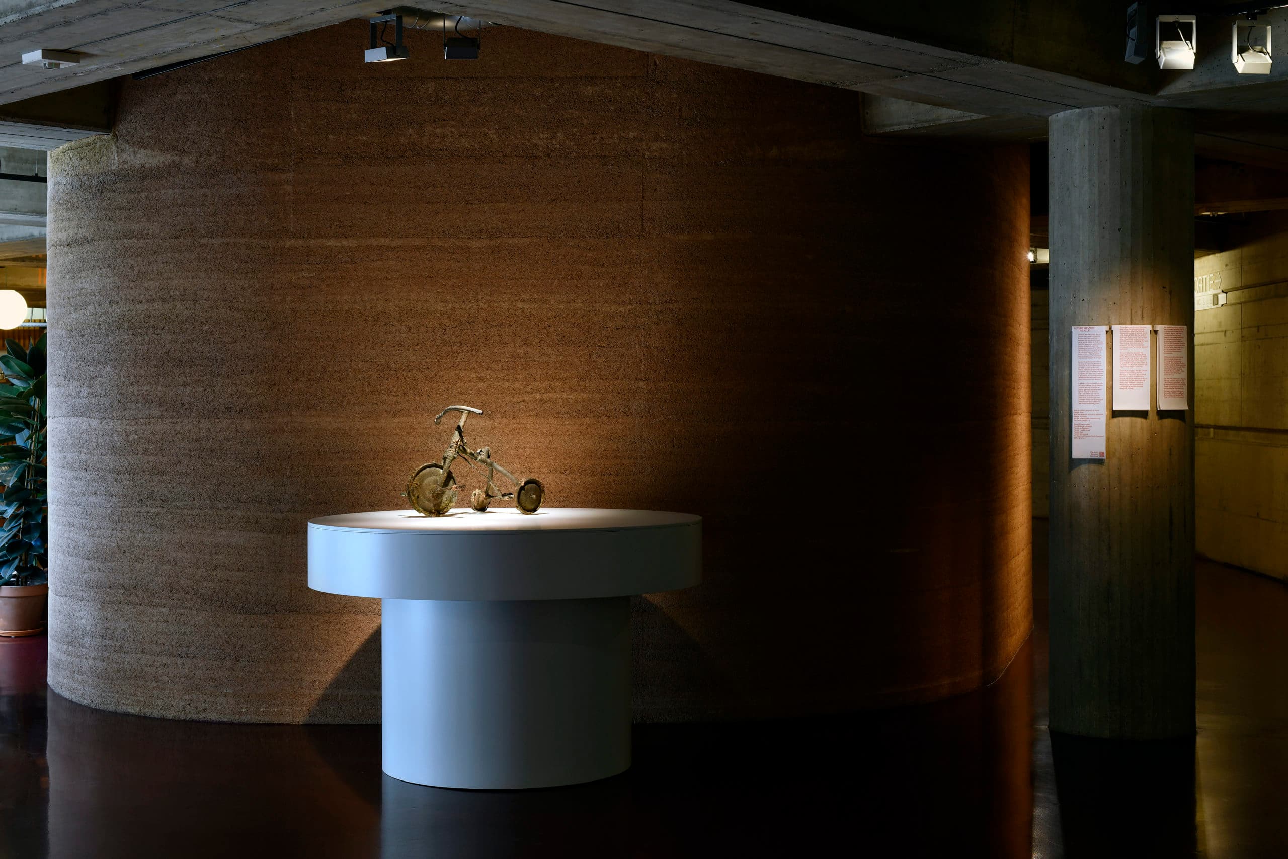 A sculpture of a metal tricycle placed on a white plinth with direct lighting, in front of a brown, textured cylindrical wall.