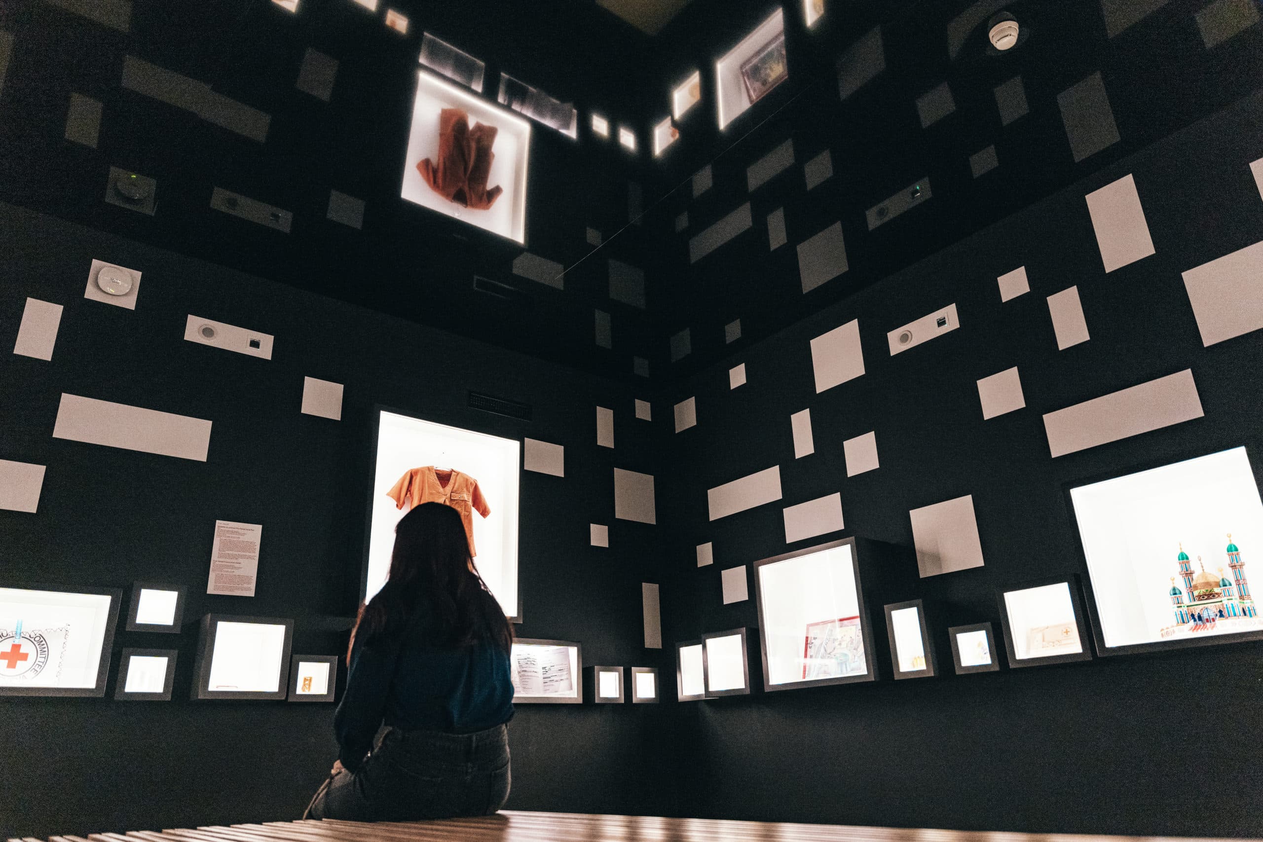 A person sitting with their back to the camera is looking at an exhibition room with dark walls, dotted with numerous frames and illuminated screens of various sizes displaying objects and illustrations.
