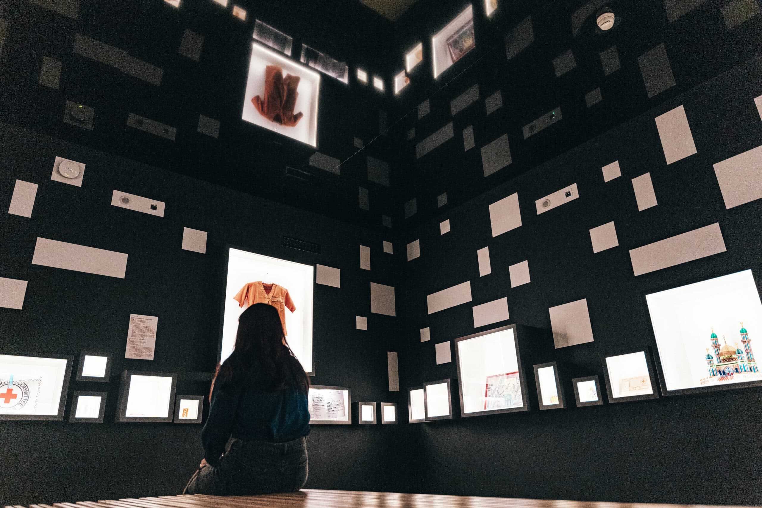 Une personne assise de dos observe une salle d’exposition aux murs sombres, dans laquelle de nombreux cadres lumineux de tailles variées présentent des objets.