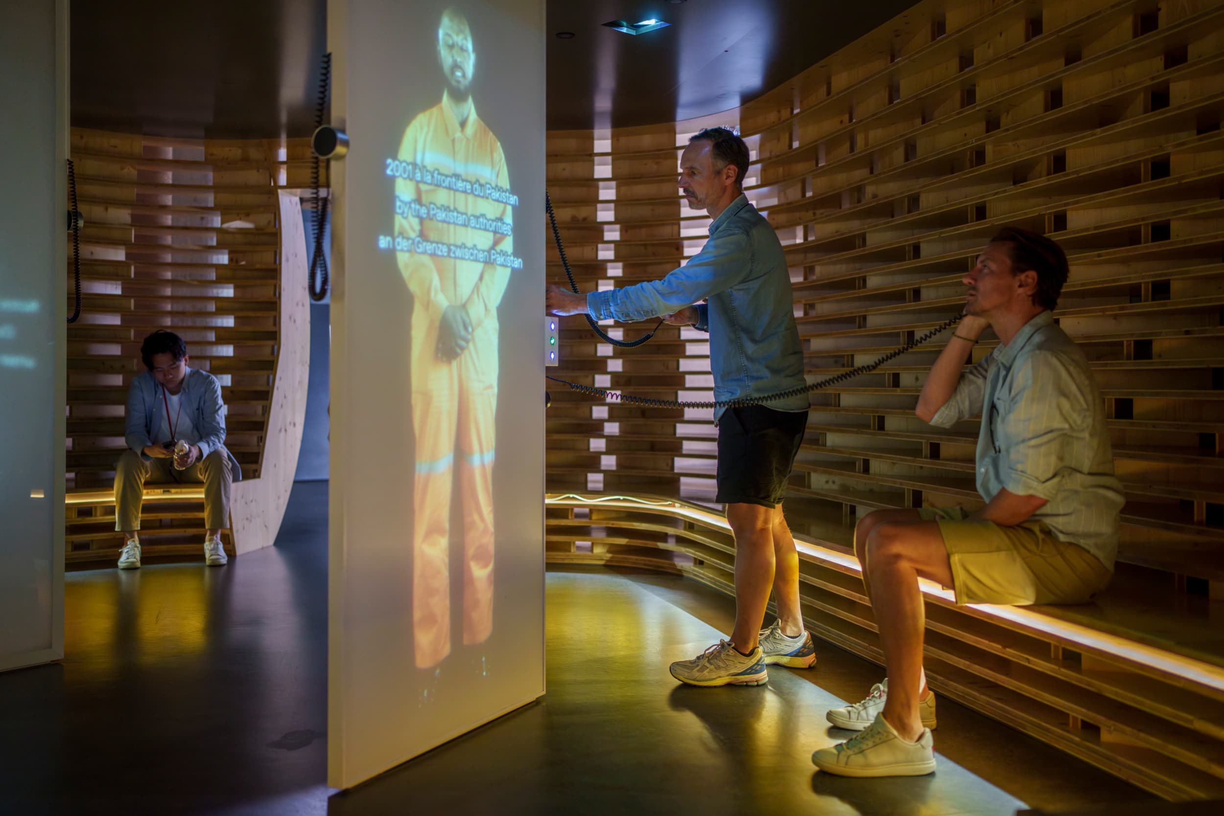 Two visitors are listening through headphones to a testimony projected onto a screen, featuring a man in an orange work uniform.
