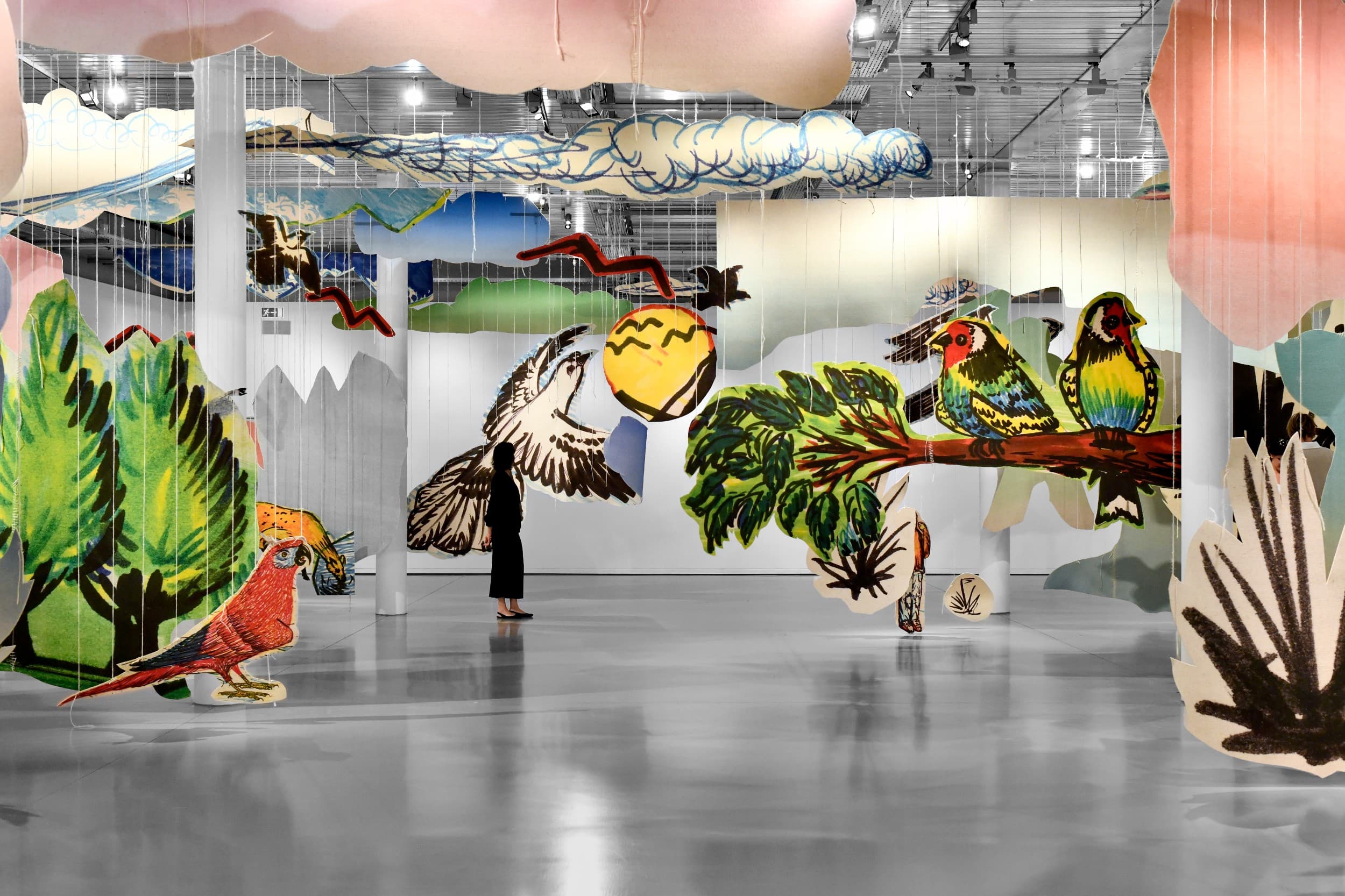 Installation artistique comportant des découpes géantes et colorées d'oiseaux, de branches et de feuillages suspendues dans l'air, des visiteurs circulent entre les découpes.