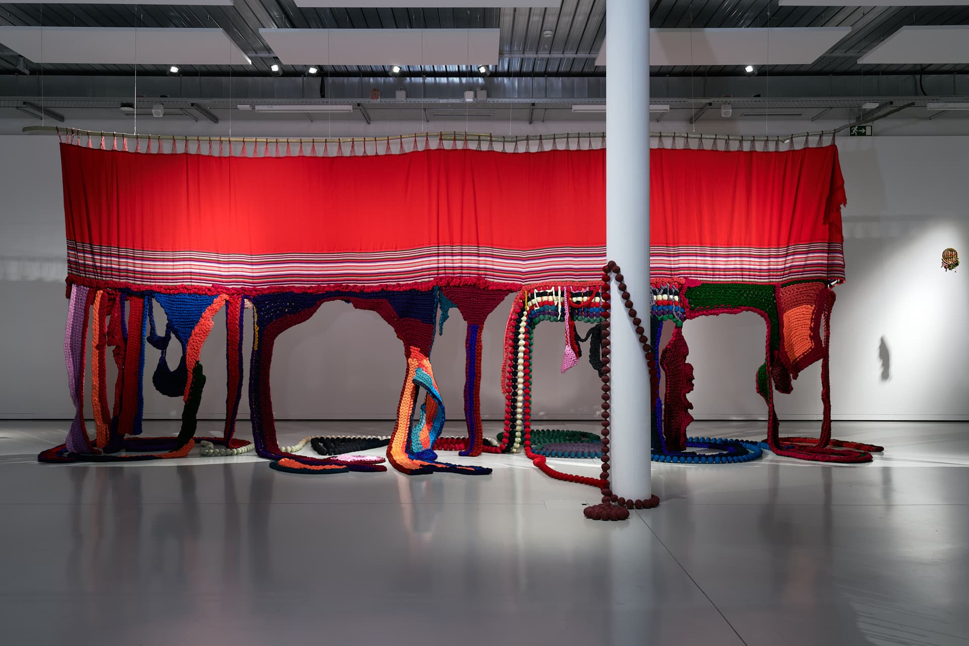 Aufnahme des Werks „Germinar desde las profundas raíces“. Eine große rote Textilinstallation ist aufgehängt, und bunte Fransen und Webelemente fallen in einem Ausstellungsraum bis zum Boden herab.