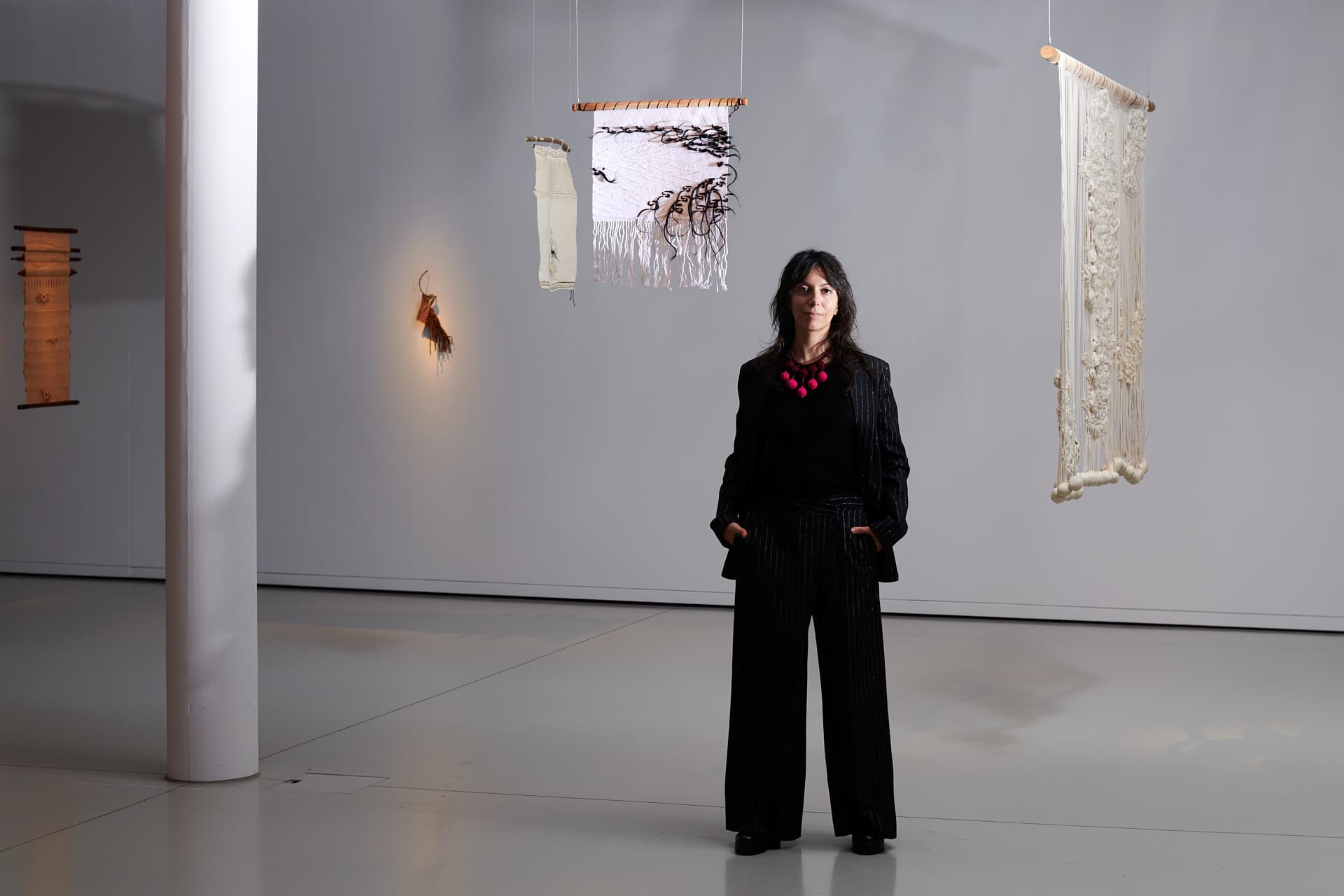 Porträt der Kuratorin Elisa Rusca. Eine schwarz gekleidete Frau steht in einem minimalistischen Ausstellungsraum, umgeben von hängenden Textilwerken in hellen Farben.