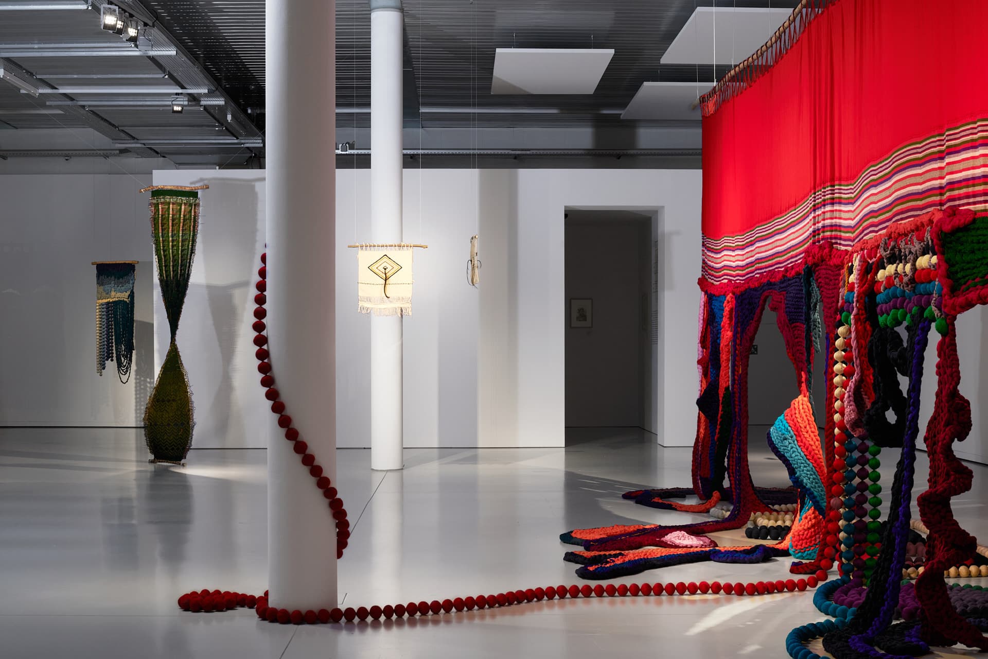 Heller Ausstellungsraum mit weißen Säulen, in dem sich eine rote und mehrfarbige Textilinstallation über den Boden und entlang einer Wand erstreckt, begleitet von weiteren hängenden Stücken im Hintergrund.