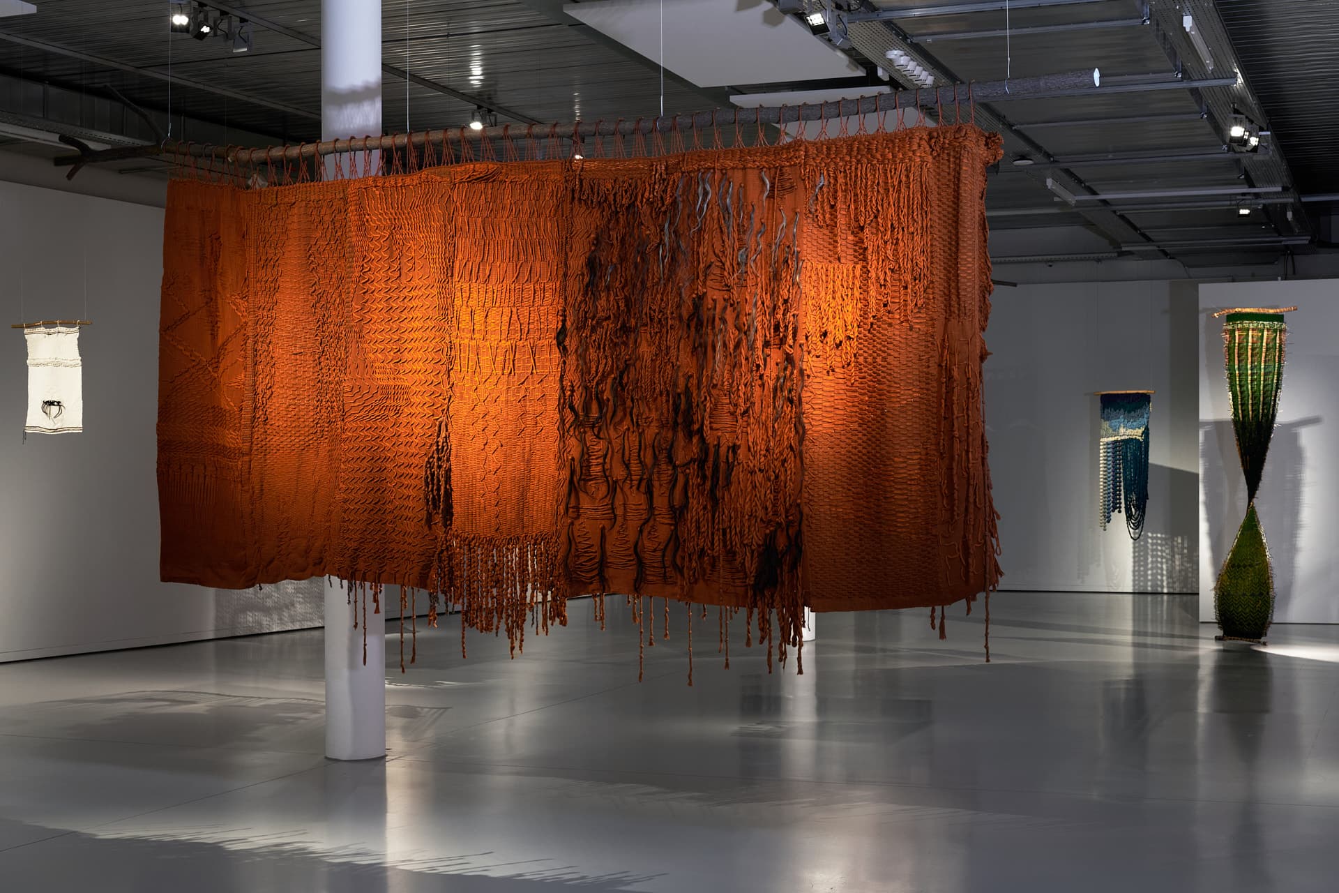 Aufnahme des Werks „Mi historia en nudos, al dorso de mi Güipil“. Eine große, braun-orangefarbene Textilinstallation, die in der Mitte einer Galerie aufgehängt ist und aus Knoten, herabfallenden Fransen sowie schwarzen und grauen Haarsträhnen besteht, beleuchtet von Strahlern.