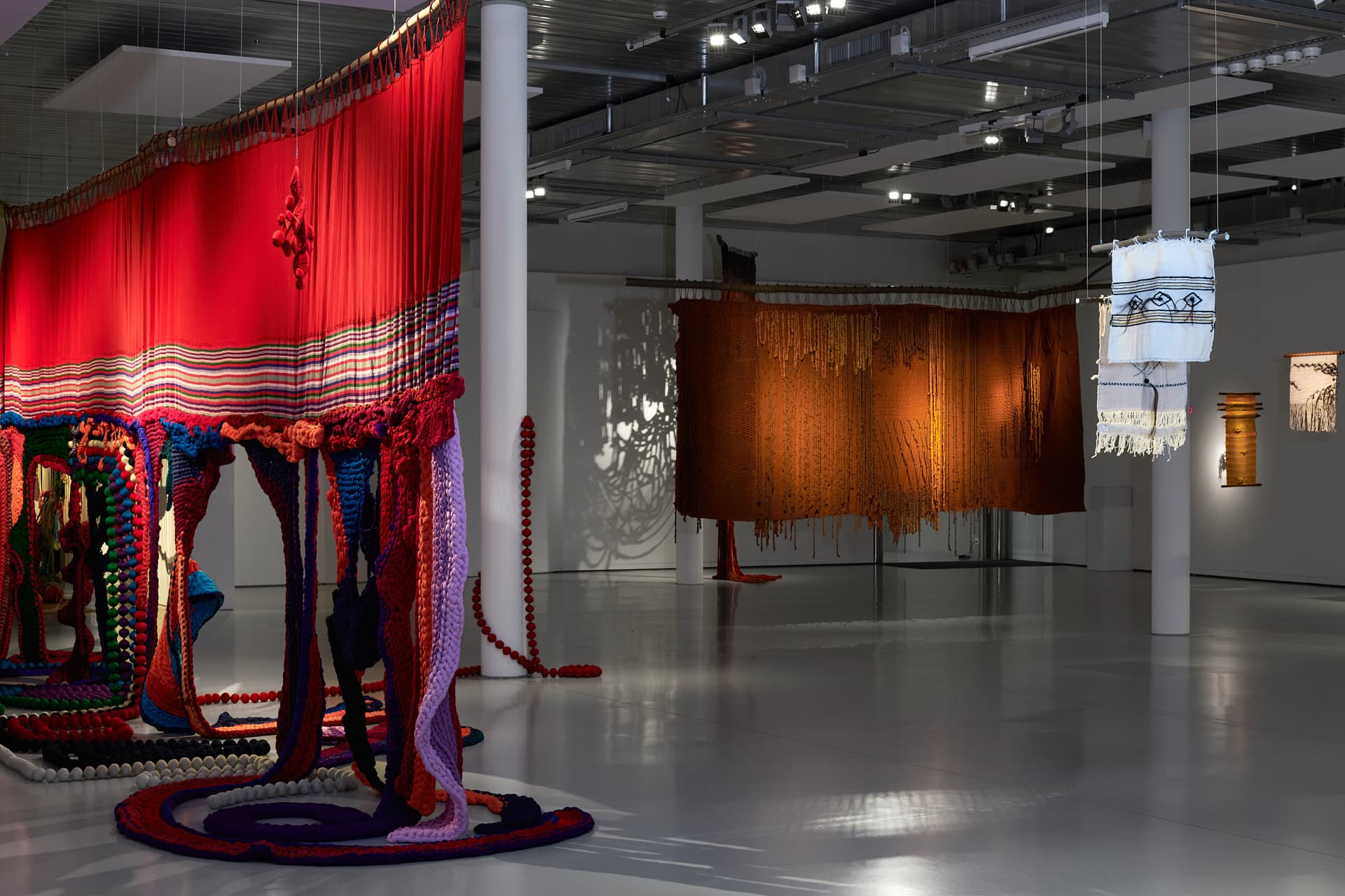 Bunte Wandteppiche in verschiedenen Größen hängen von der Decke eines Ausstellungsraums.