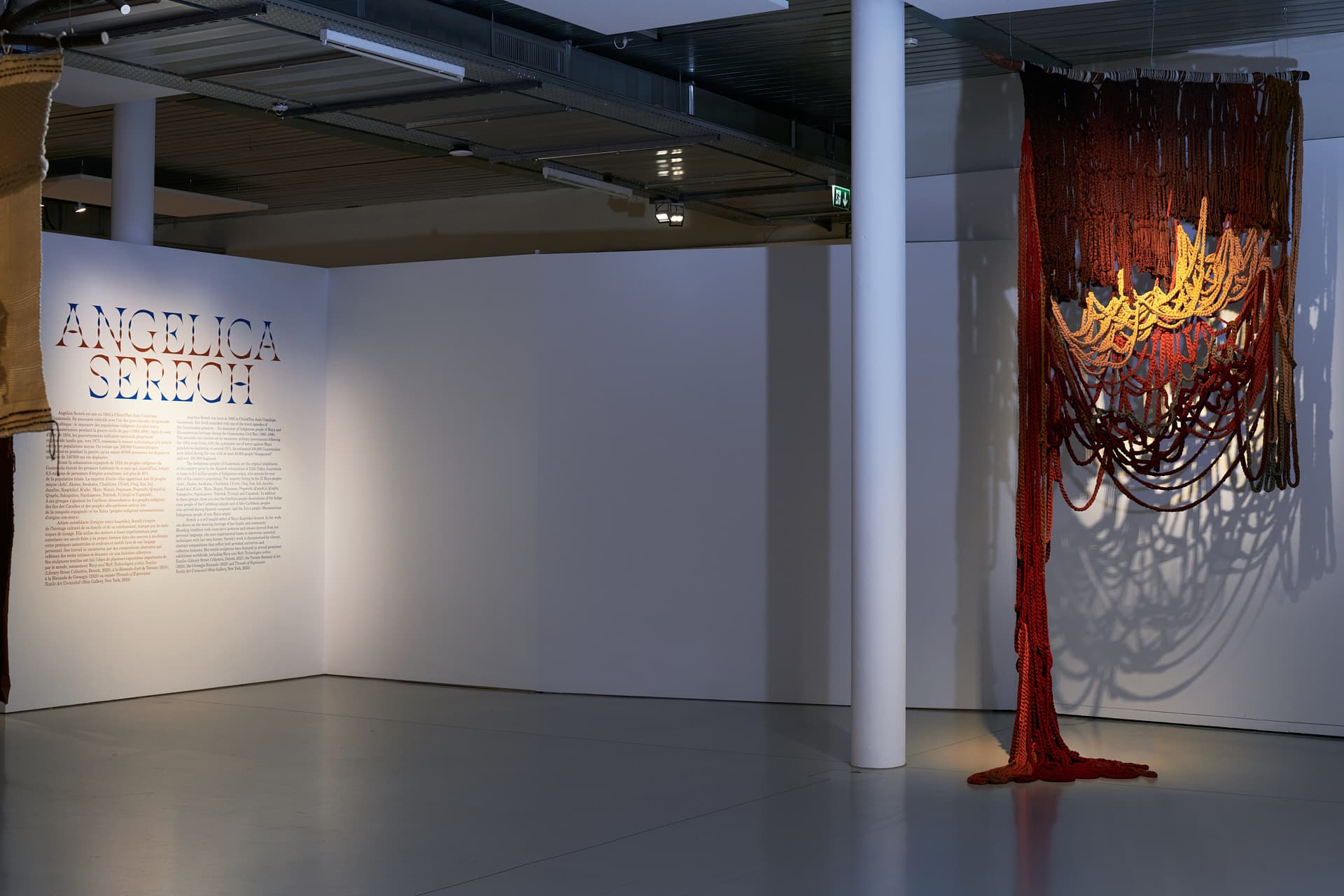 Aufnahme des Werks „Sororidad“, eines ocker- und beigefarbenen Wandteppichs mit ockerfarbenen Flechtwerken, die bis zum Boden reichen. Links eine weiße Wand mit einer Beschreibung der Ausstellung und dem Namen der Künstlerin.