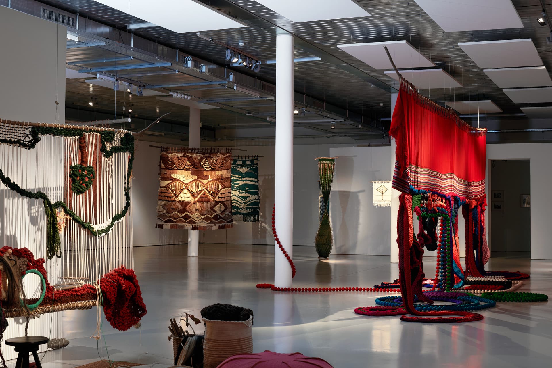 Gesamtansicht eines Ausstellungsraums mit mehreren farbenfrohen Textilinstallationen, die von der Decke hängen und Werke mit geometrischen Mustern sowie Elemente in leuchtenden Farben wie Rot und Grün umfassen.