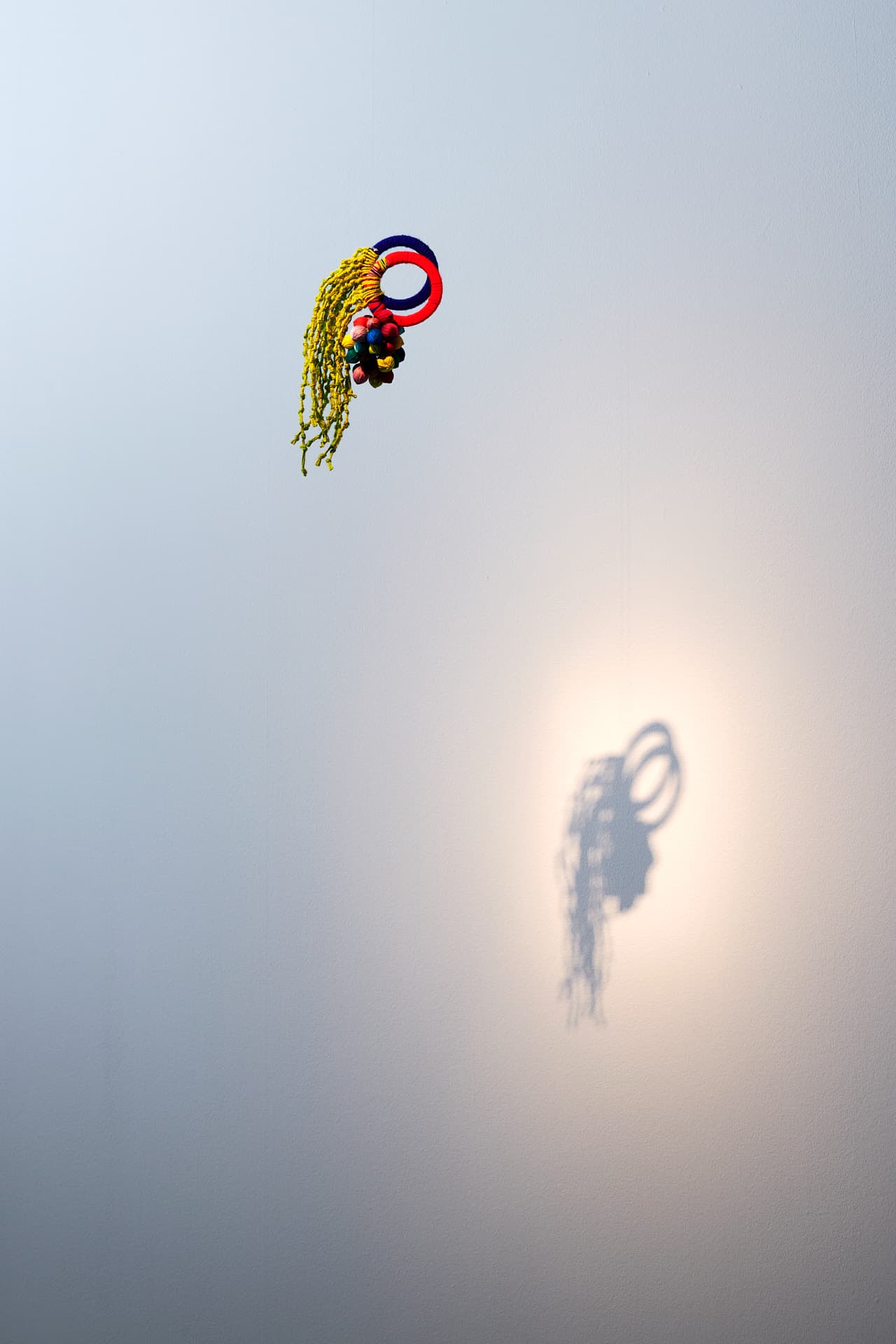 Aufnahme von „Euforia en Chixot / Comalapa“. Hängende Textilinstallation, bestehend aus zwei roten und blauen Kreisformen, an denen Fäden mit Knoten und kleinen Knäueln befestigt sind, die einen Schatten auf eine helle Wand werfen.