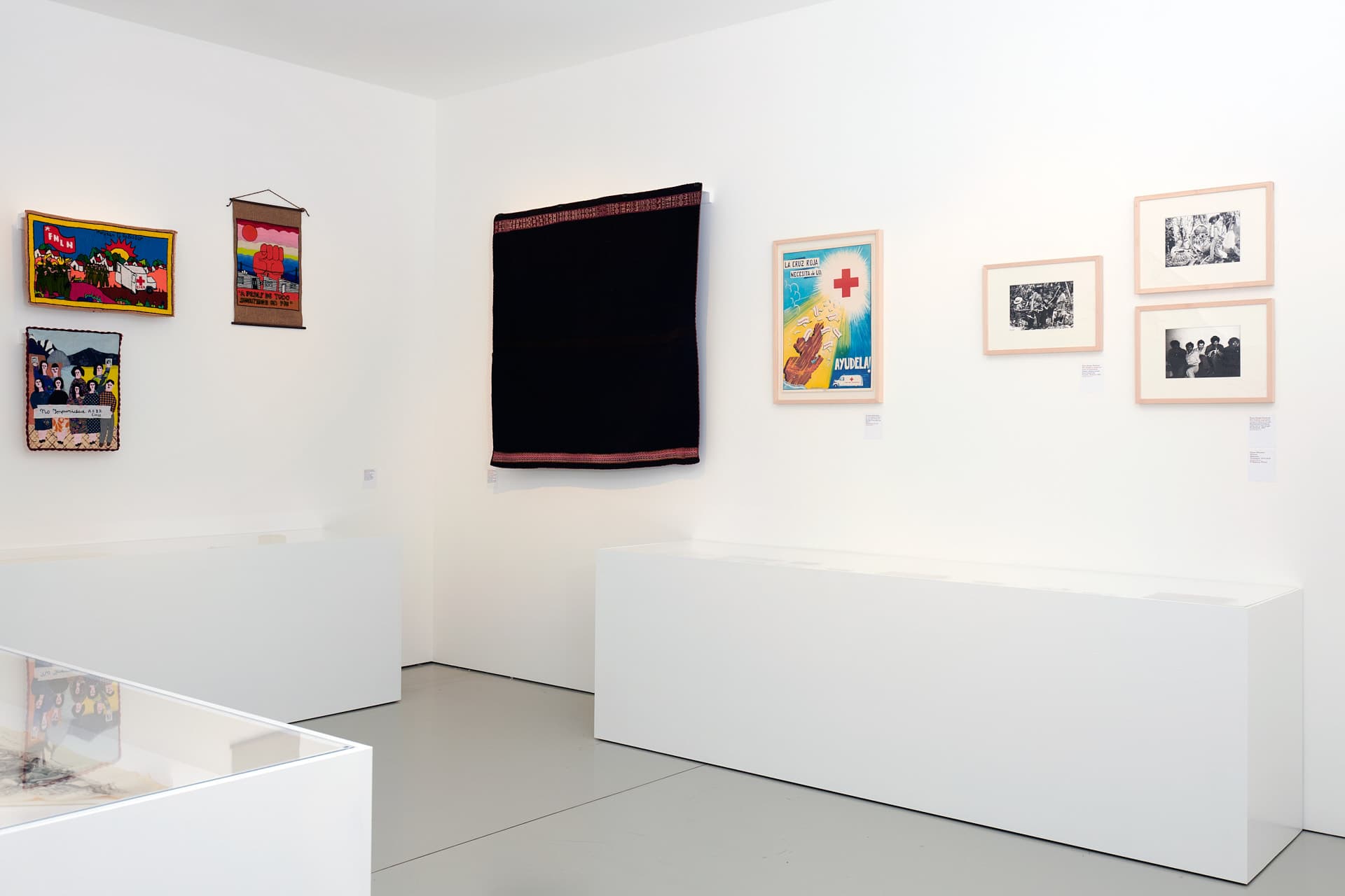 Ausstellungsraum mit weißen Wänden, in dem ein dunkler Wandteppich, Schwarz-Weiß-Fotografien und ein Plakat zu sehen sind. An der linken Wand hängen drei kleine, farbenfrohe Textilwerke.
