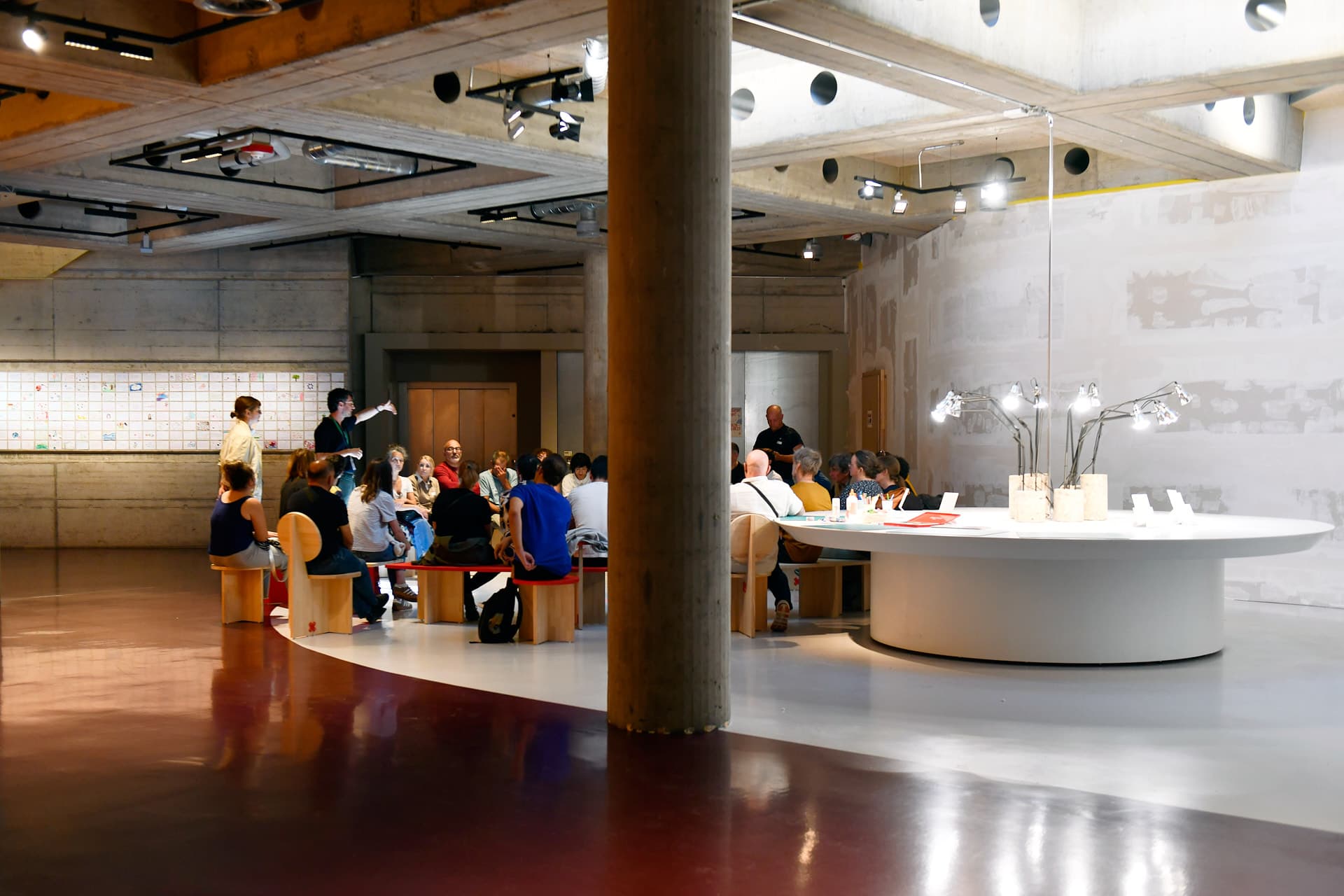 A group of people sitting in a circle in a brightly lit basement space. Behind them, a large, white, circular table with numerous lamps in the centre.