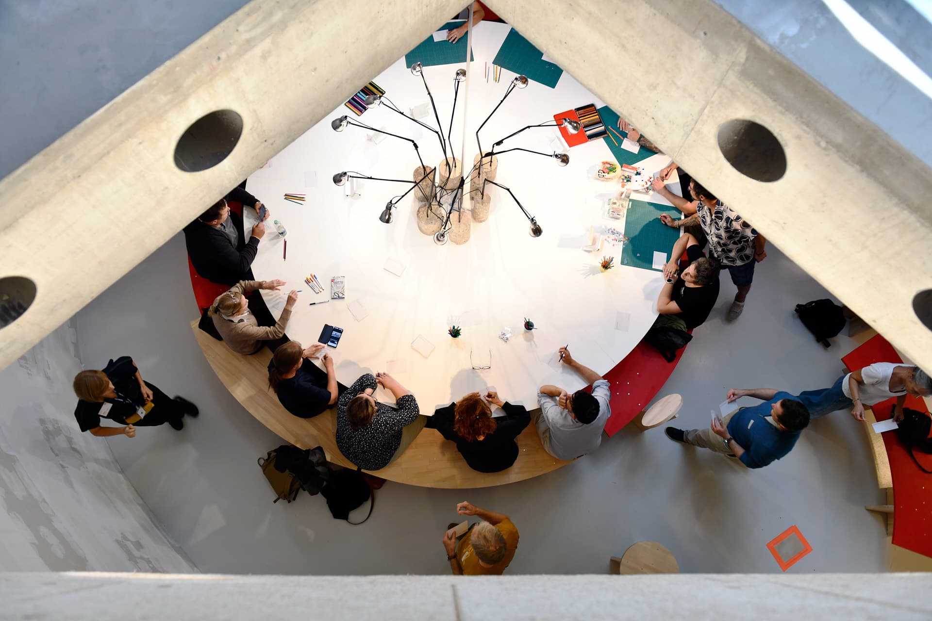 Vue de dessus de personnes assises autour d'une grande table blanche aménagée avec du matériel créatif et des lampes articulées.