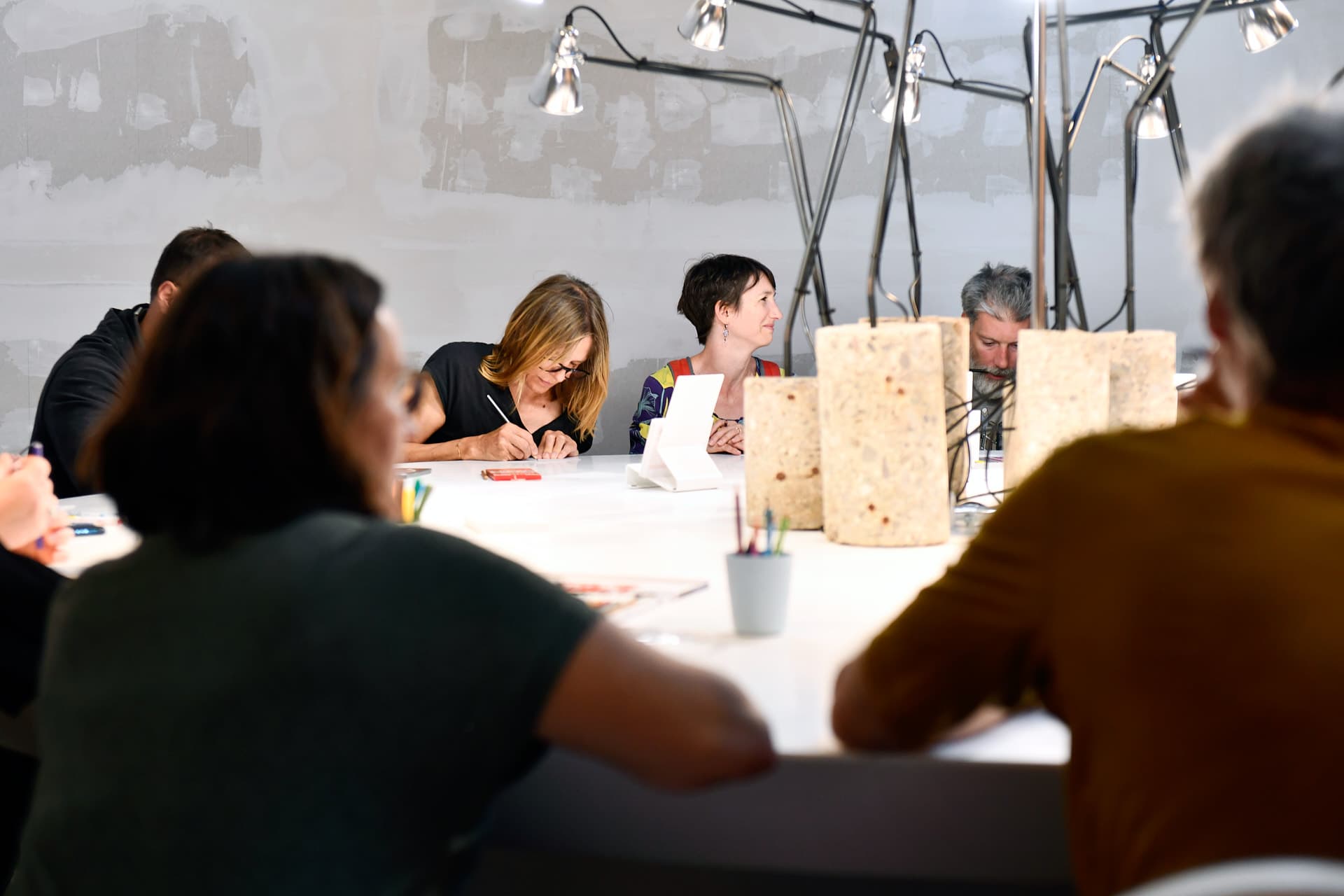 Personnes assises autour d'une table blanche en train de dessiner avec du matériel de dessin et des lampes au centre de la table. Au premier plan, deux silhouettes à contre-jour de personnes assises autour de cette table.