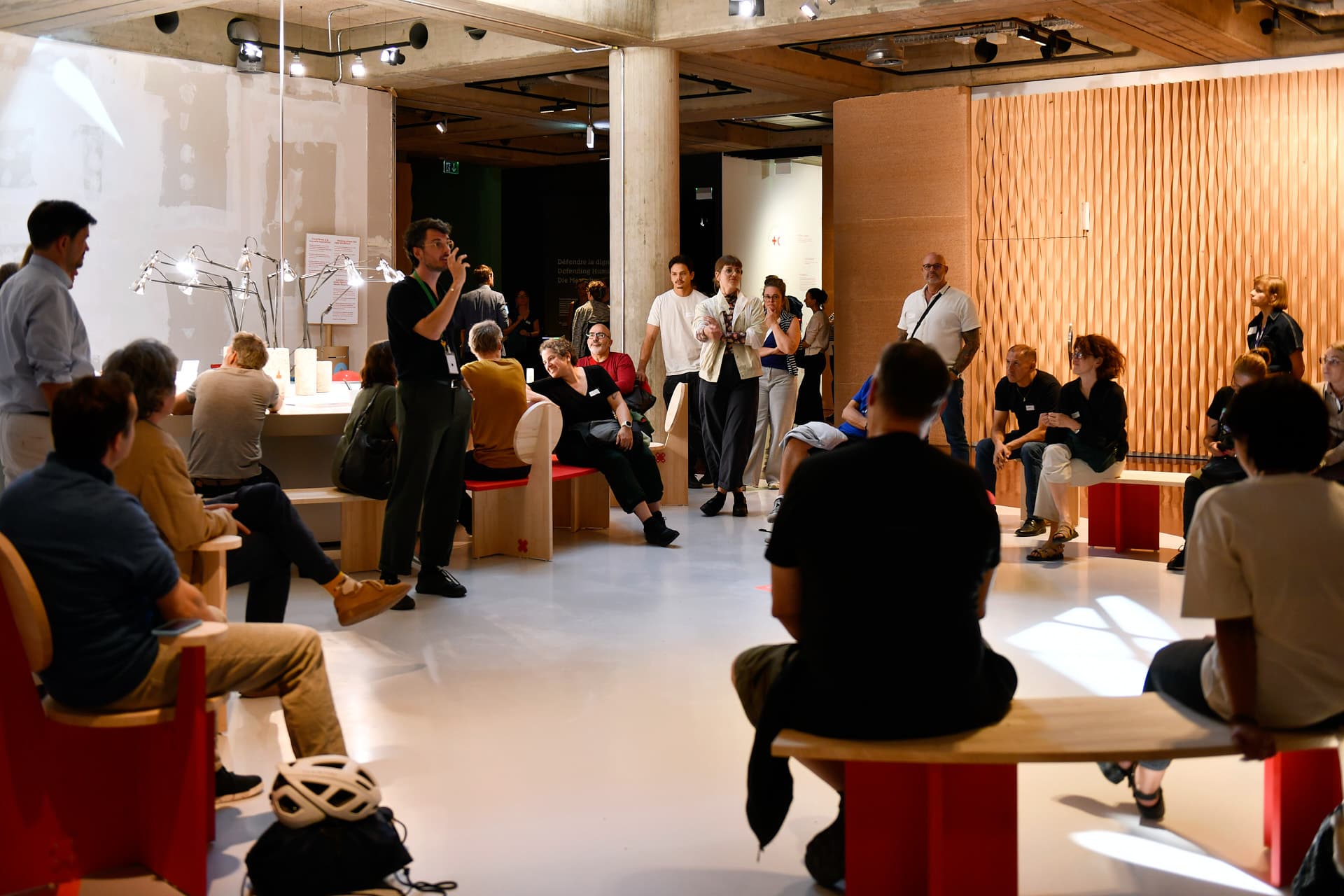 Groupe de personnes réunies dans un espace contemporain, certaines assises et d’autres debout, tandis qu’un intervenant prend la parole au centre de la salle dans une ambiance chaleureuse.
