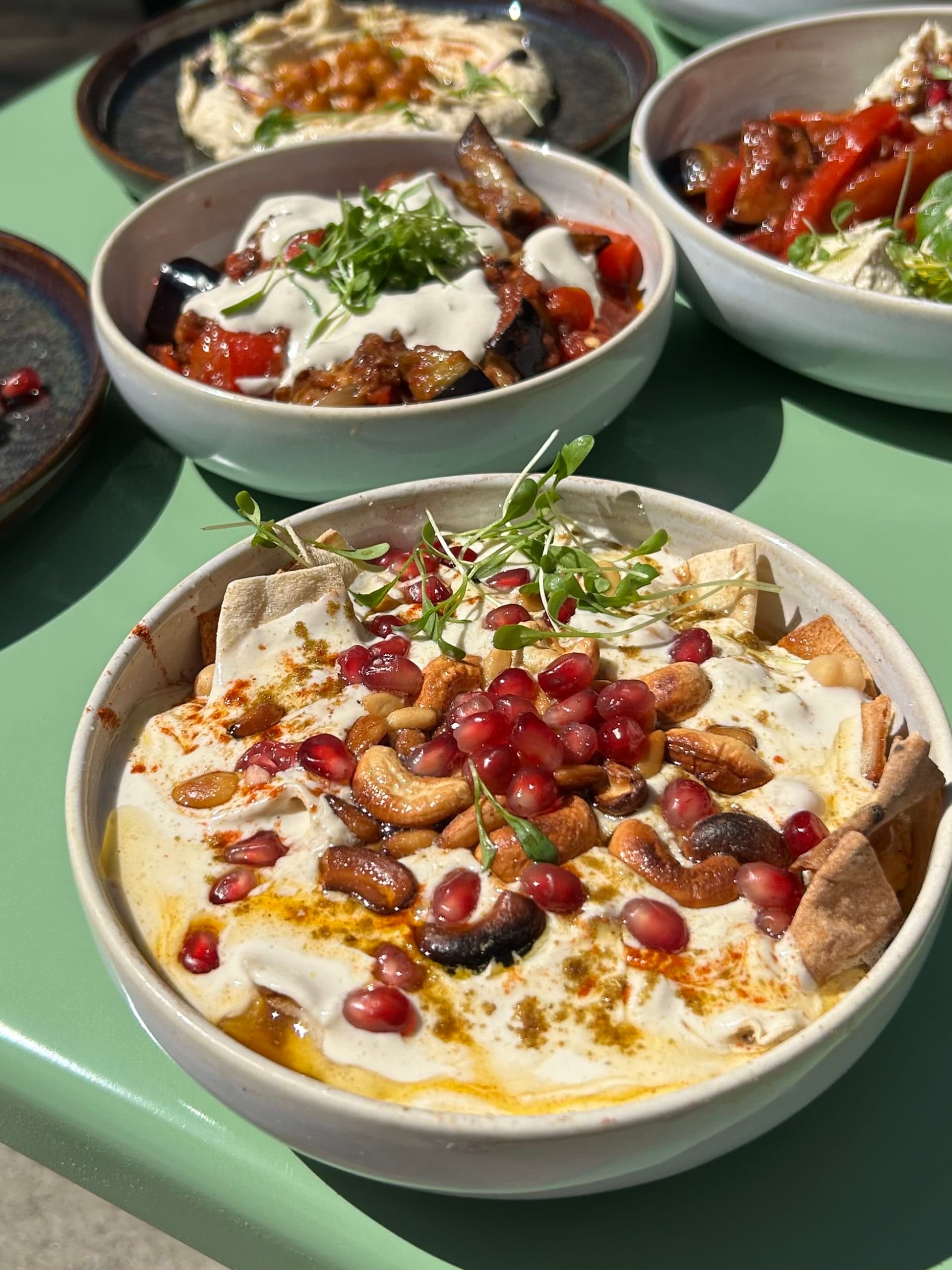 Bol de houmous garni de pois chiches, tomates cerises, herbes fraîches et huile d'olive posé sur une table au soleil, dans le fond d'autres plats sont visibles.