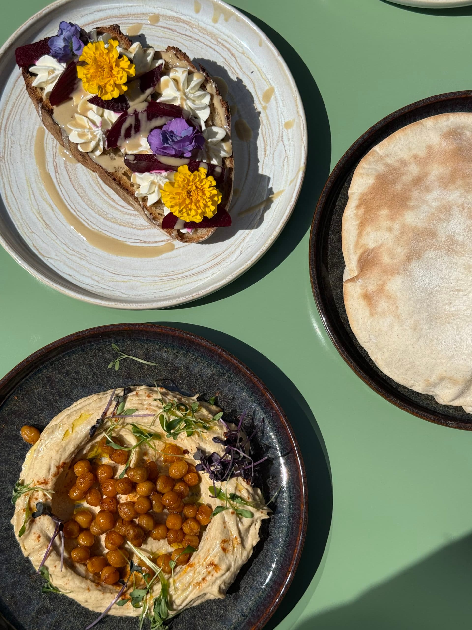 Deux assiettes de cuisine méditerranéenne, prises depuis le dessus, l'une contenant du houmous avec des pois chiches grillés au centre et l'autre, une tartine de fromages frais avec des fleurs violettes et jaunes comme décoration.
