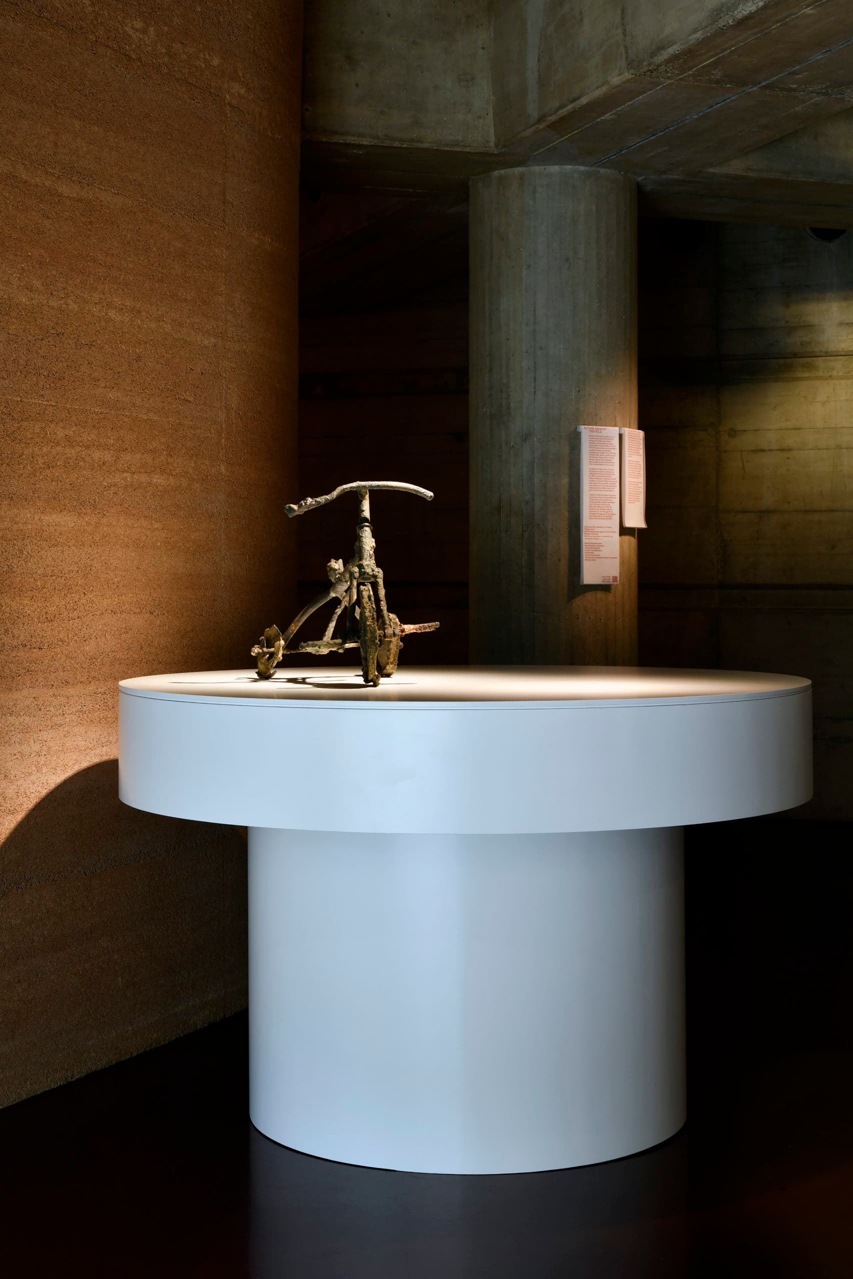 Skulptur eines Dreirads aus Metall, ausgestellt auf einem breiten, runden, weißen Sockel, der von einem Scheinwerfer angestrahlt wird. Im Hintergrund eine strukturierte braune Wand und eine Betonsäule, auf der sich eine Erläuterungstafel befindet.
