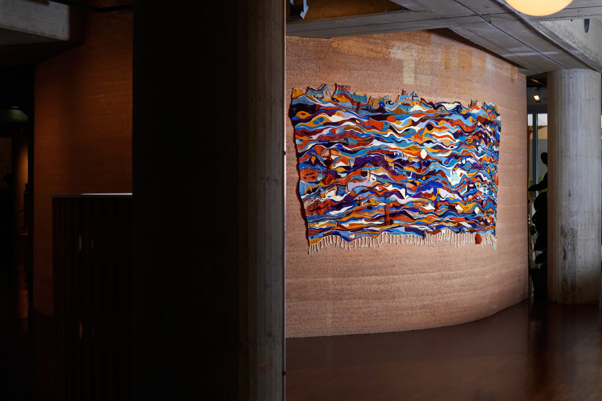 Tapisserie murale colorée aux motifs ondulés abstraits, accrochée sur un mur texturé en chanvre.