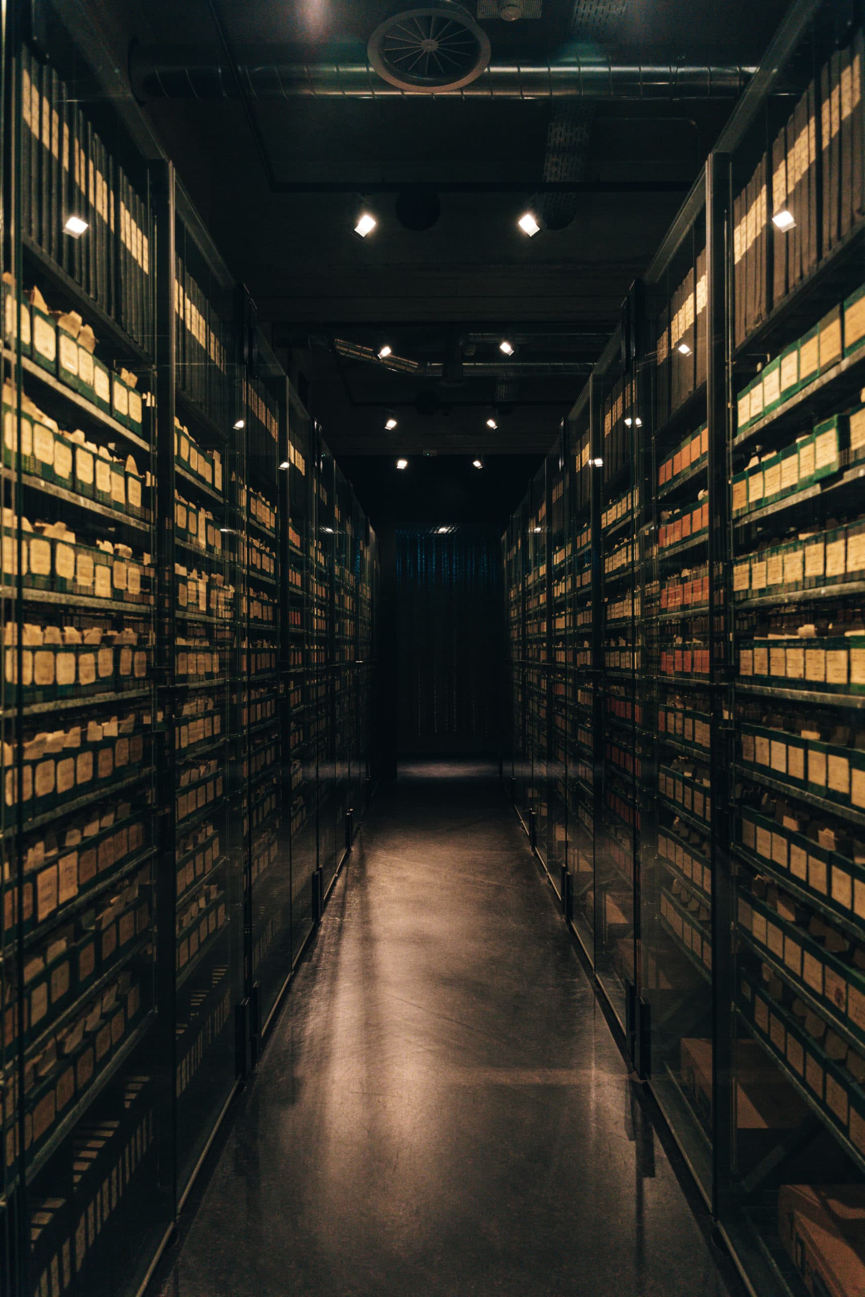 Ein schmaler Gang, gesäumt von hohen, verglasten Regalen, die mit beschrifteten Archivkisten gefüllt sind und in gedämpftem Licht stehen.
