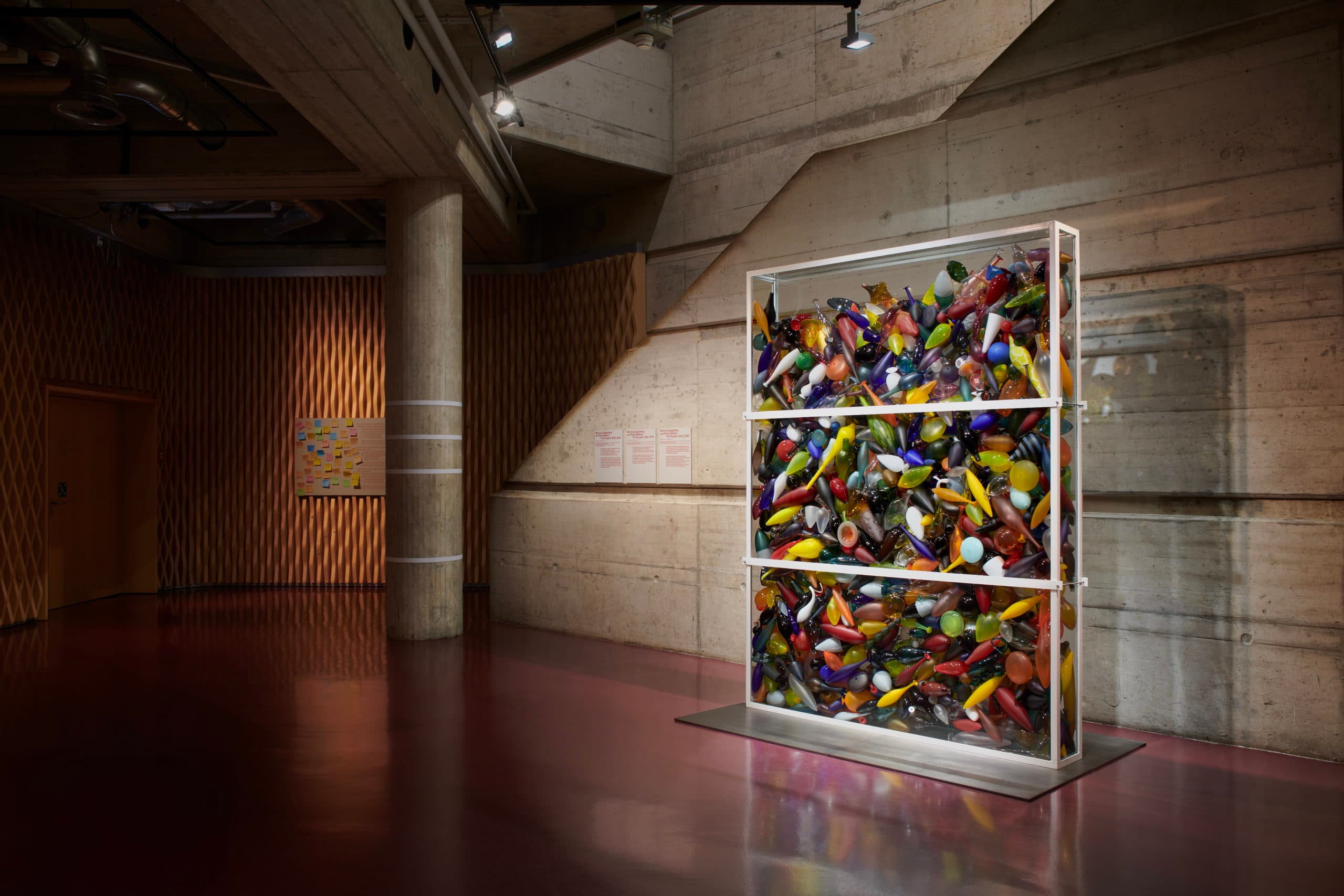 Une grande structure vitrée rectangulaire. encadrée de métal blanc, est remplie de centaines d’objets colorés en verre, exposée devant un mur en béton et éclairé par un projecteur dans une salle tamisée.