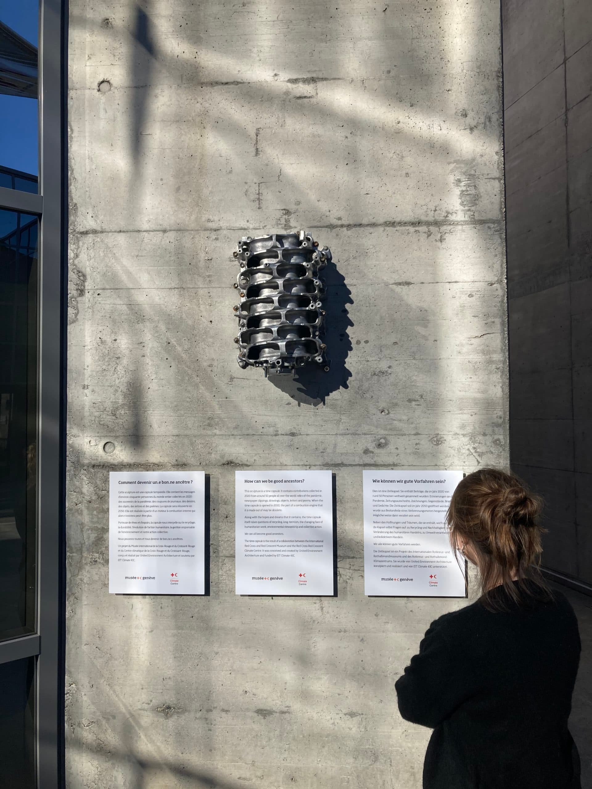 Visiteuse lisant des panneaux d’exposition sous une sculpture métallique ressemblant à un moteur de voiture fixée à un mur en béton.