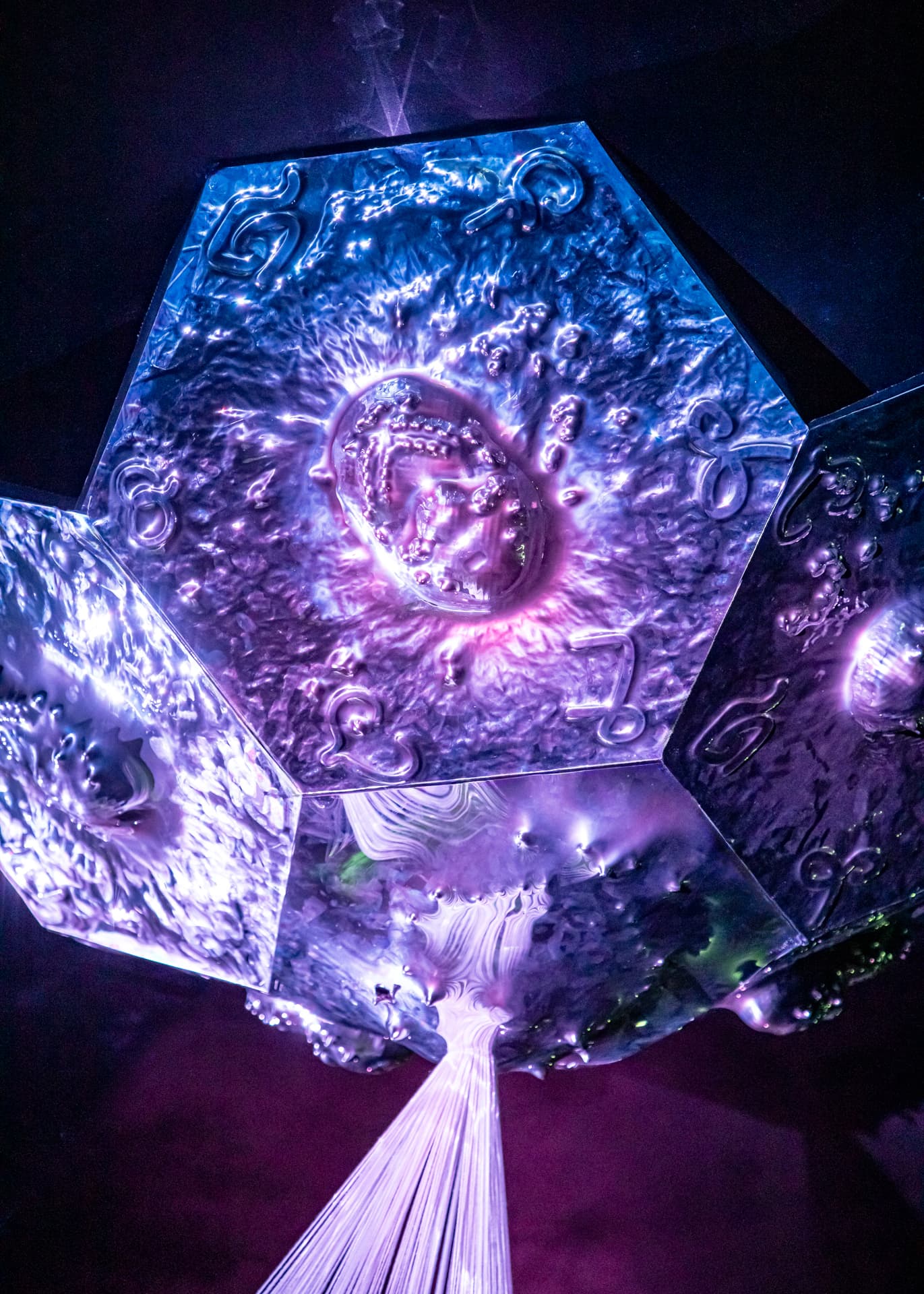 Objet géométrique hexagonal aux reflets bleutés et violets, orné de motifs gravés et reflétant une lumière violette, avec des fibres lumineuses s’en échappant vers le bas.