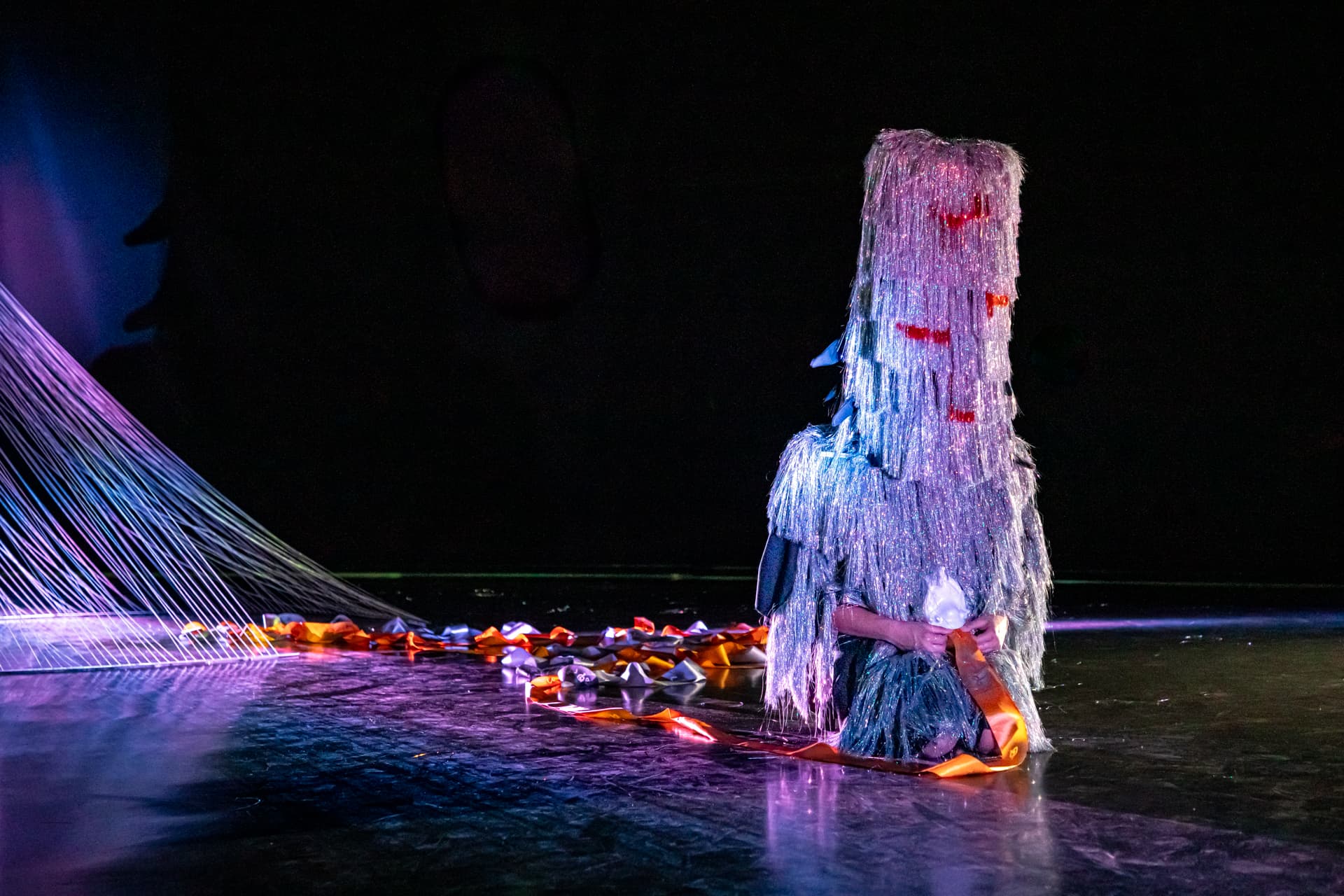 Eine Person sitzt auf der Bühne, gekleidet in einen strukturierten, glitzernden Anzug, mit einer Maske, die das Gesicht bedeckt, umgeben von bunten Bändern und einem violetten Lichtstrahl im Hintergrund.