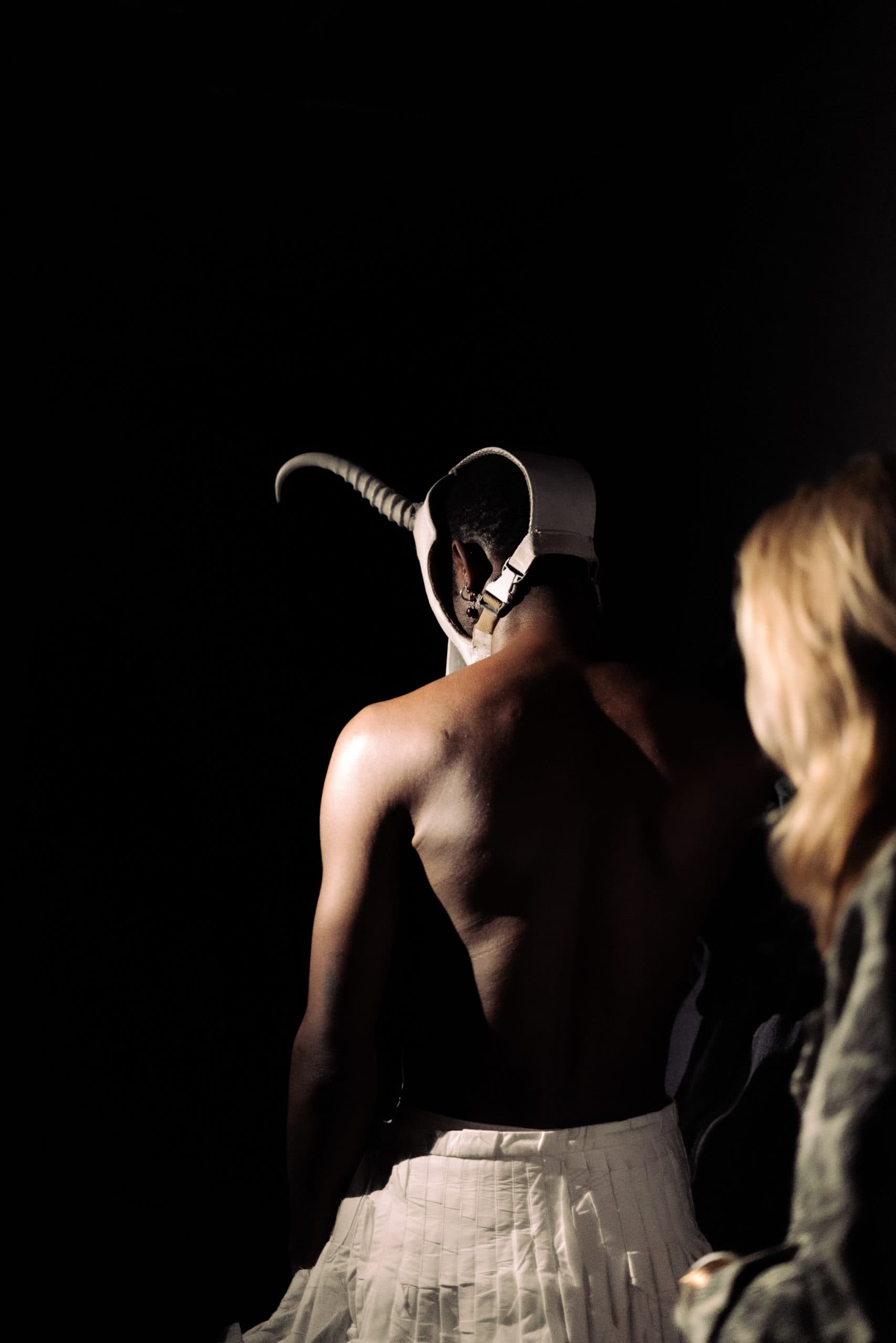 Vue arrière d’une personne portant un masque blanc avec une corne recourbée, éclairée par un spot dans l’obscurité, en compagnie d’une autre personne aux cheveux clairs.