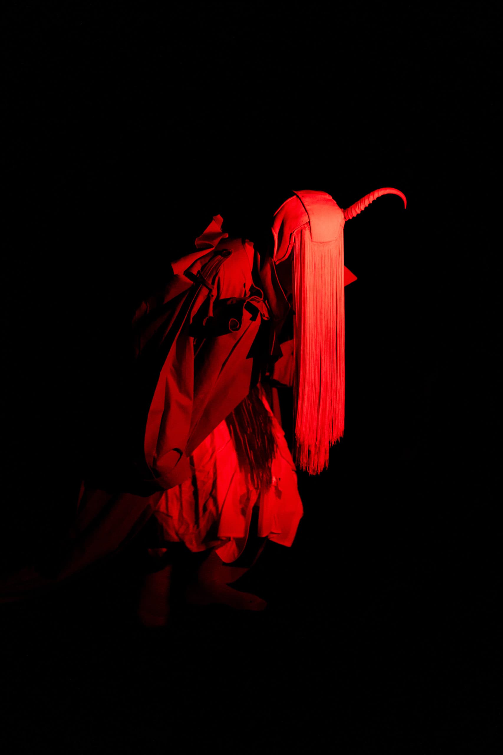Silhouette of a person wearing a red costume, wearing a mask with horns and long fringes, lit by a bright red spotlight against a black background.