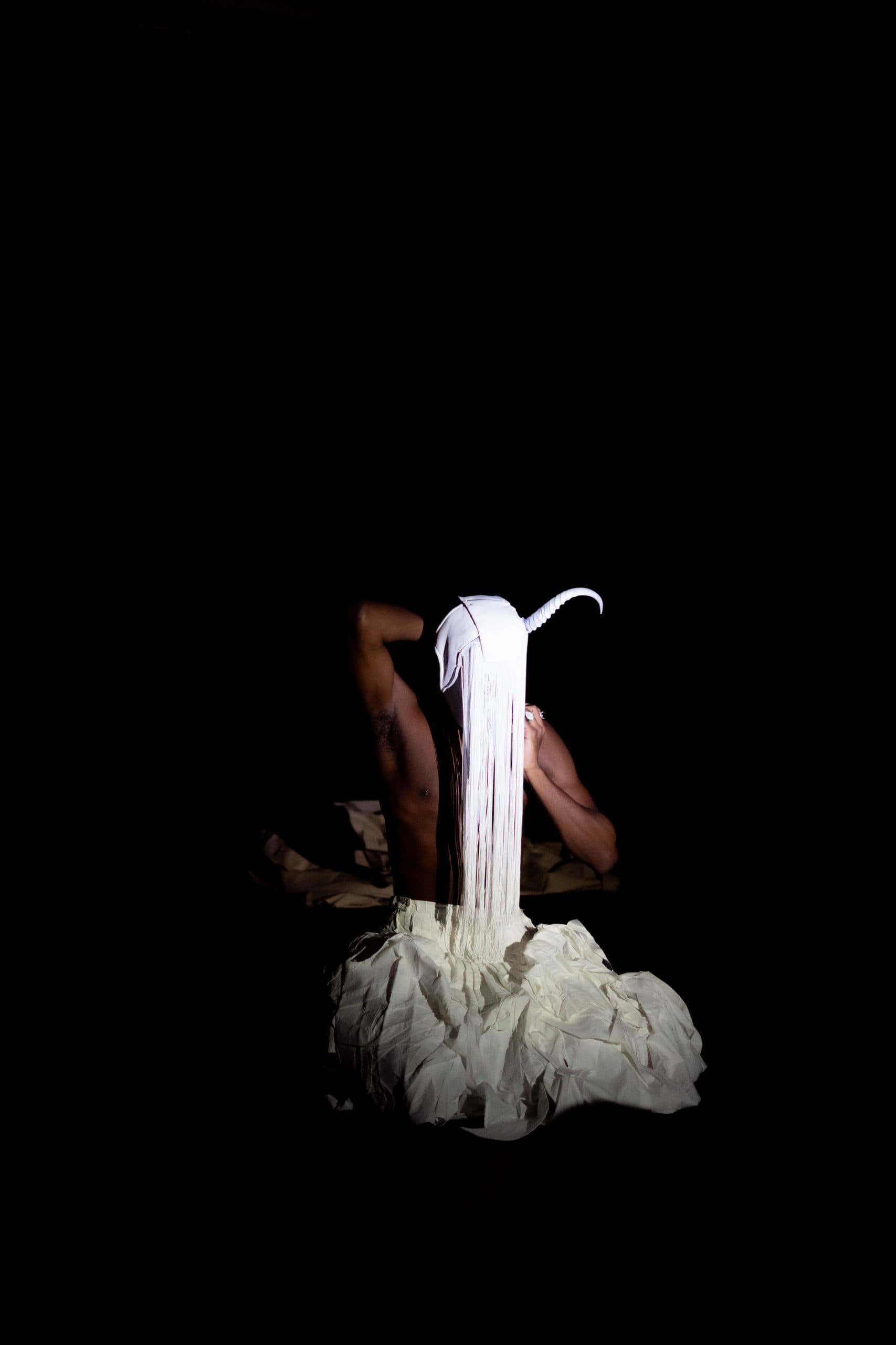 Personne assise, portant un masque blanc avec une corne et des franges, vêtue d’une jupe texturée, les mains posées sur la tête, dans une scène sombre et éclairée par un spot.