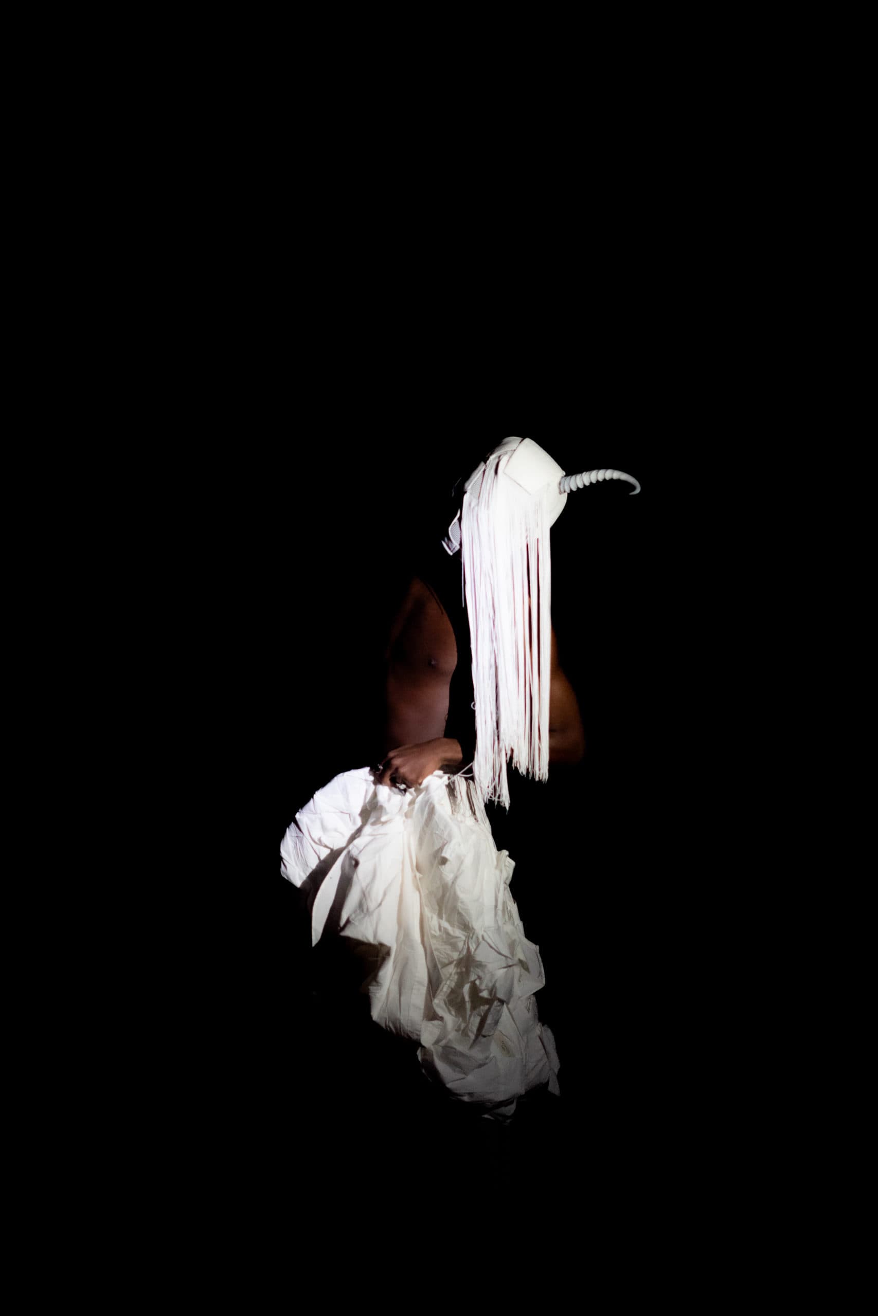 Person wearing a white mask with a curved horn and fringes, dressed in a loose, crumpled gown, lit by a spotlight against a black background.