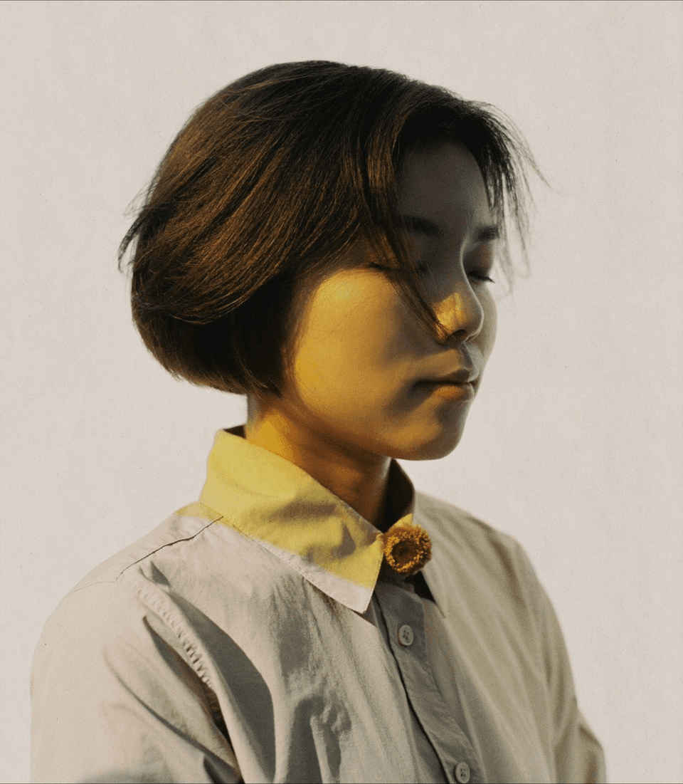 Portrait of a short, dark-haired woman posing in three-quarter profile against a light-coloured wall with her eyes closed. A yellow-tinted light reflects on her skin.
