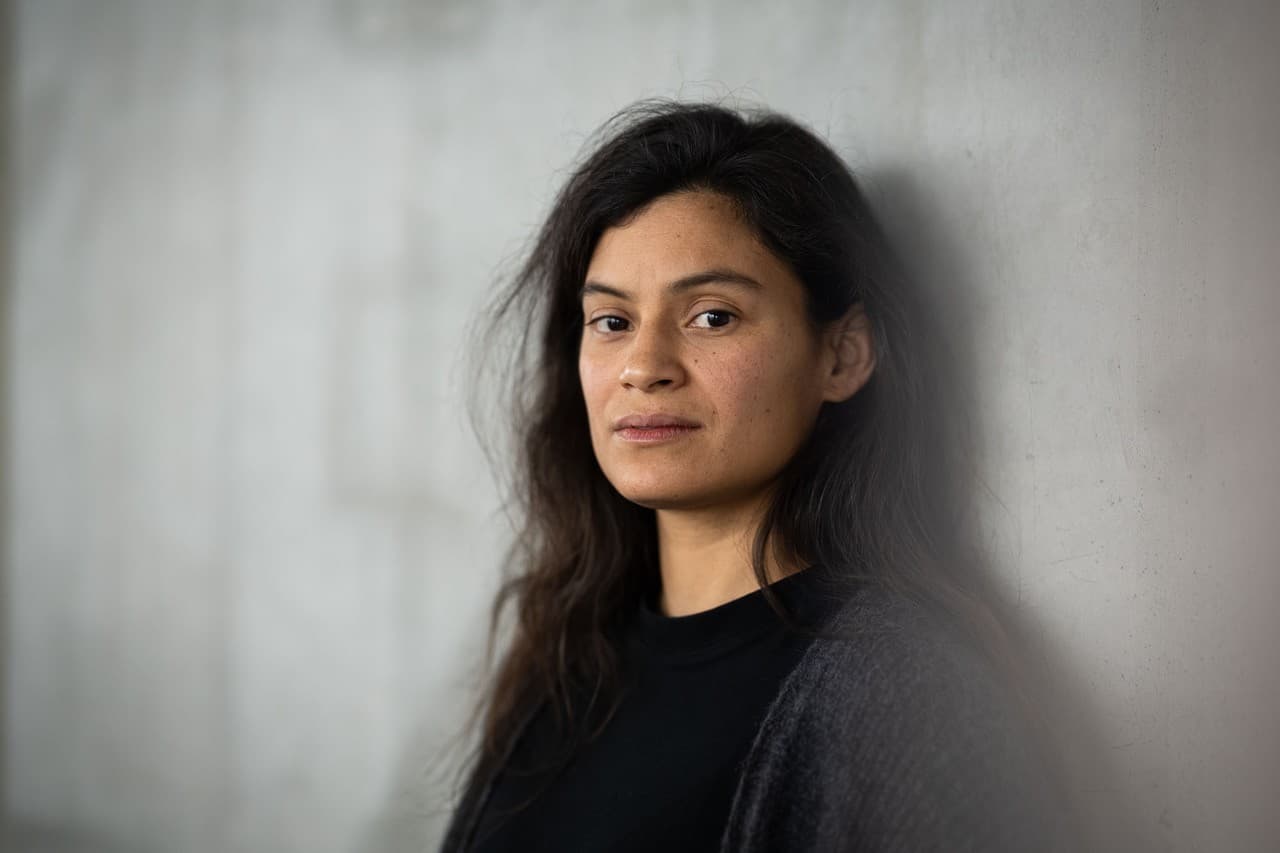 Une femme adossée contre un mus gris en béton regarde l'objectif et sourit légèrement.