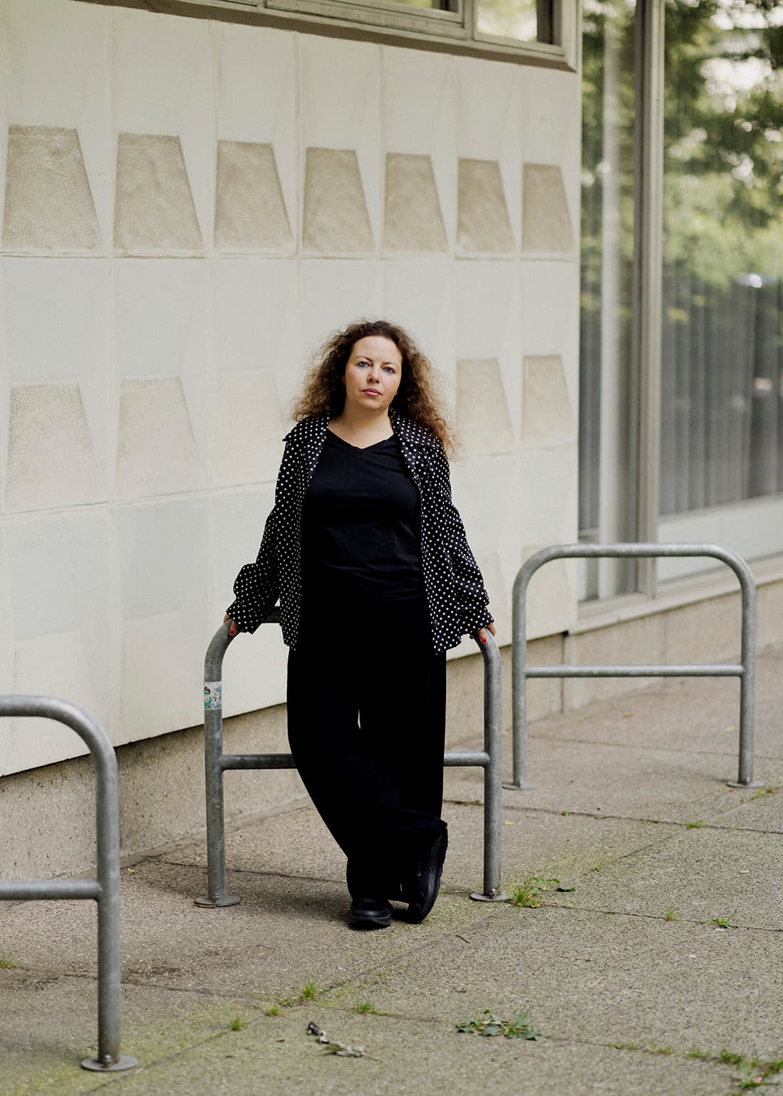 Frau mit lockigem Haar, die zwischen zwei Metallreifen auf einem Bürgersteig steht, sich an eine helle Fassade lehnt, schwarz gekleidet ist und eine gepunktete Jacke trägt, in die Kamera schaut.
