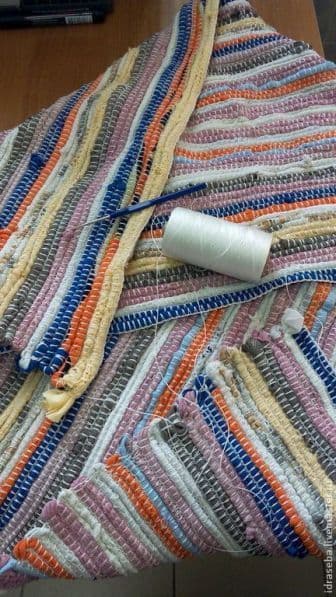 Gestreifter, mehrfarbiger Webteppich in Arbeit, mit einer weißen Garnrolle und einer Häkelnadel darauf.