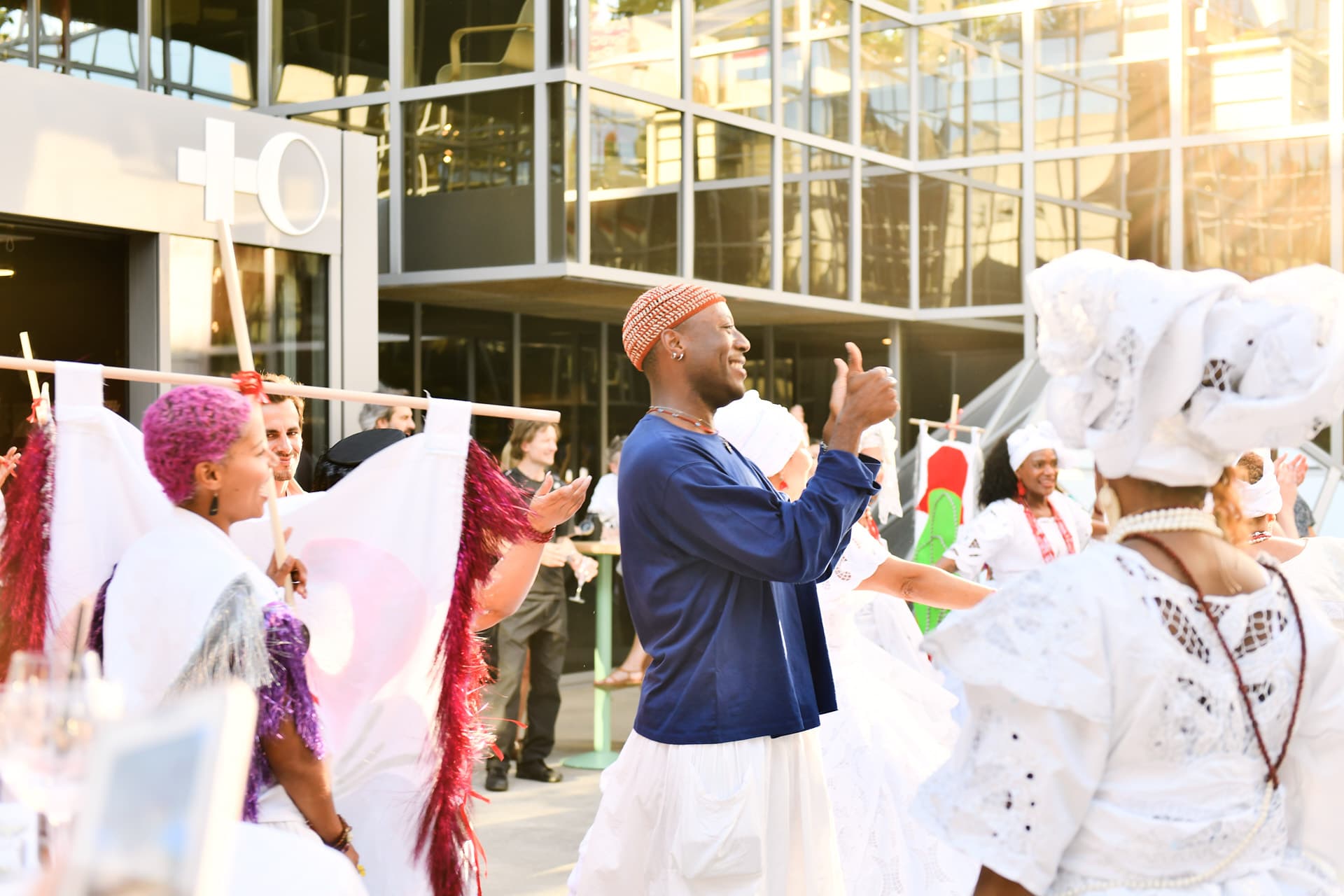 Des participants en tenues traditionnelles blanches et colorées dansent et applaudissent devant un bâtiment vitré lors d'une célébration culturelle en extérieur.