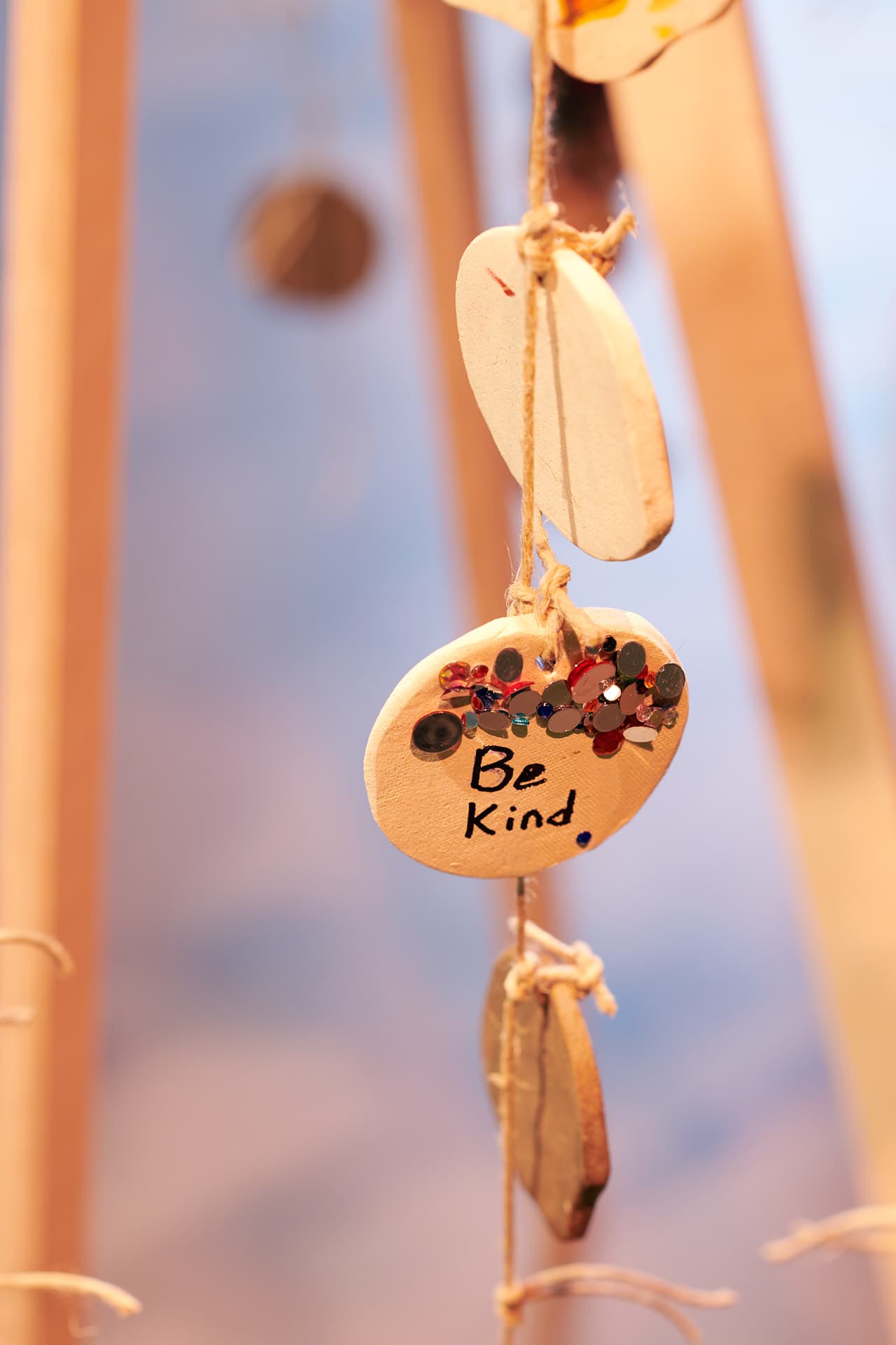 A circular ceramic charm suspended from a string alongside other ceramic circles, decorated with coloured rhinestones and bearing the inscription ‘Be Kind’ in English.