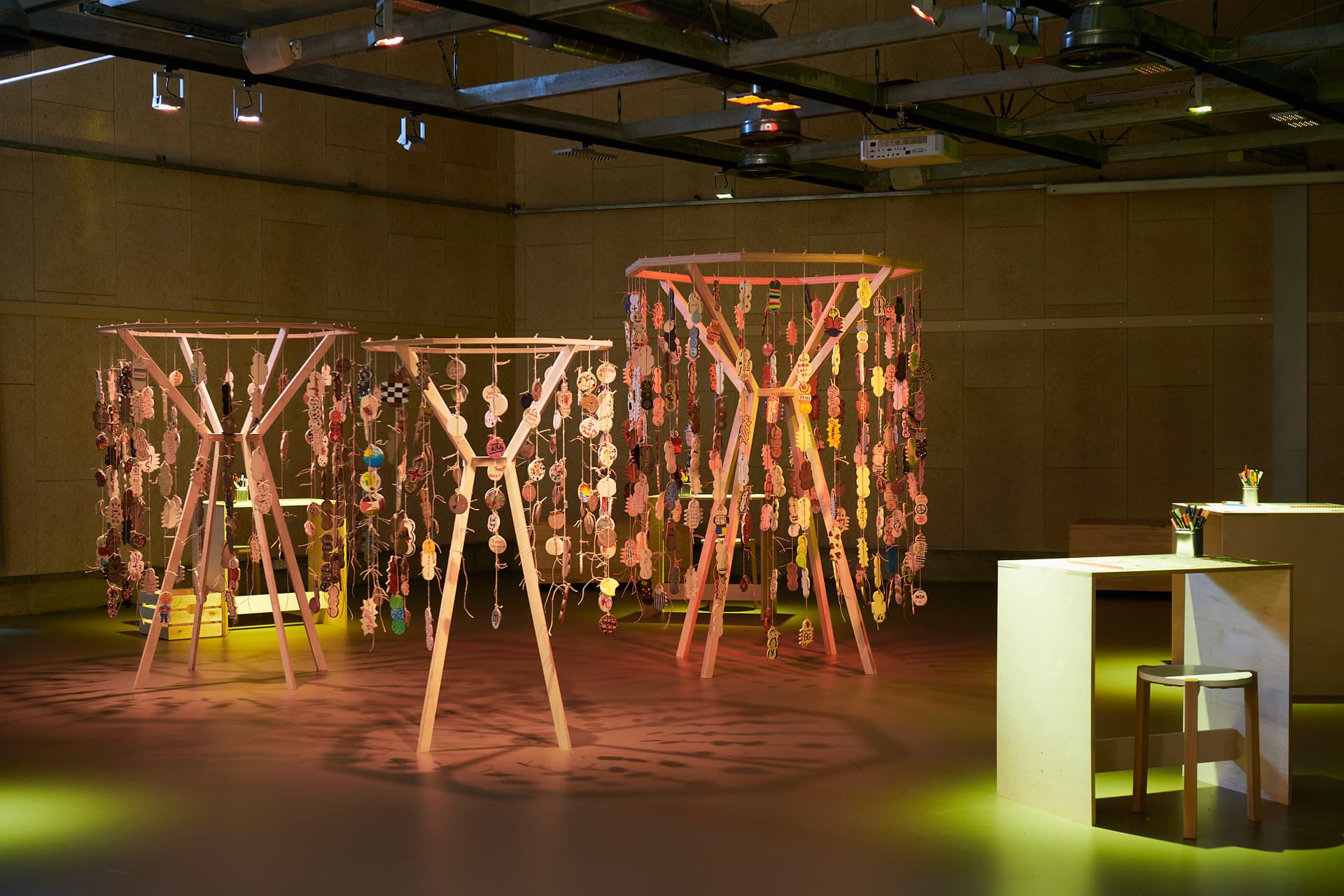 Installation artistique contemporaine dans un espace d’exposition, avec des structures en bois auxquelles sont suspendues des amulettes en céramique, disposées sur le sol et éclairées par des spots de couleur chaude.
