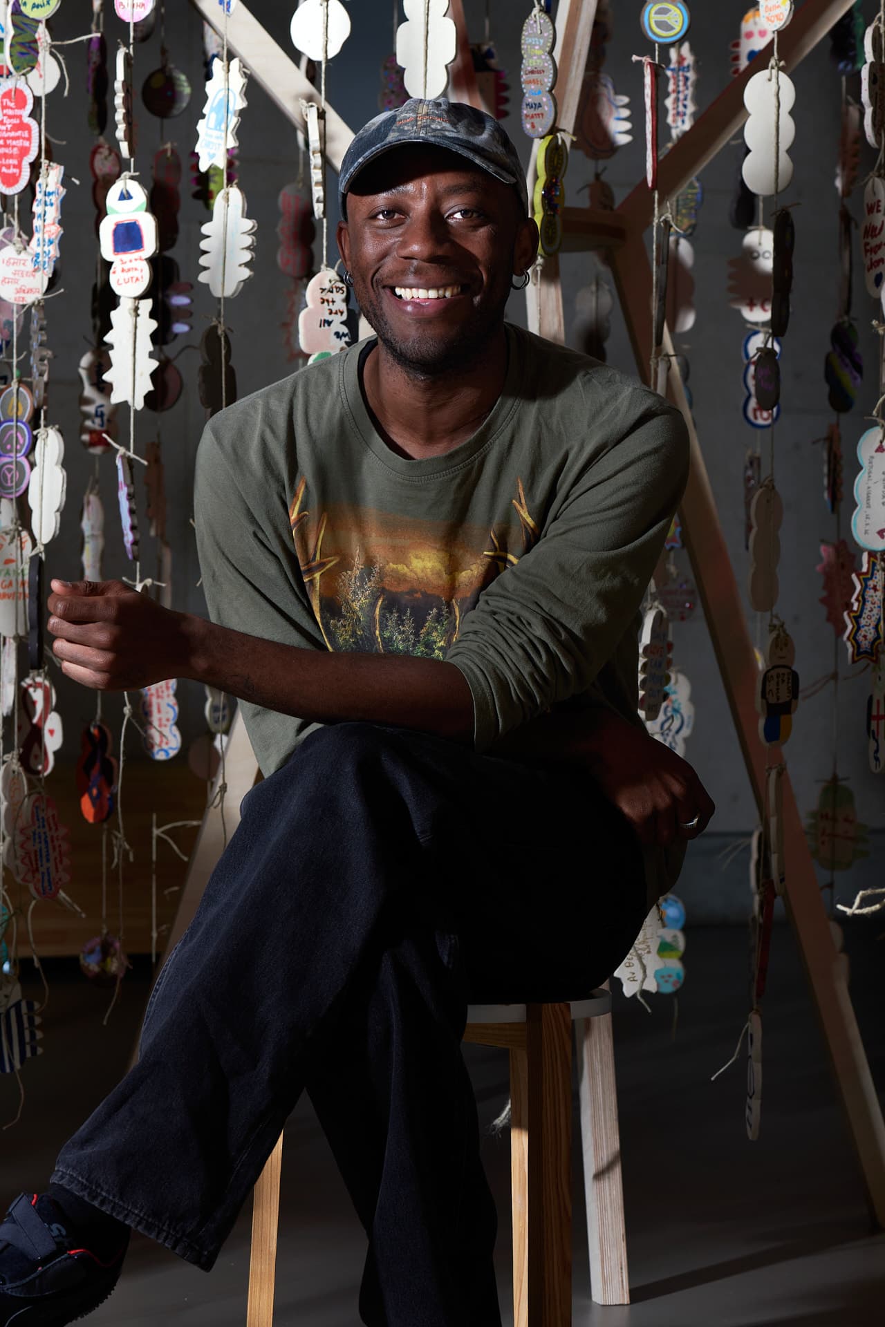 A smiling man seated on a stool inside an art installation, surrounded by hanging ceramic amulets, looking relaxed.