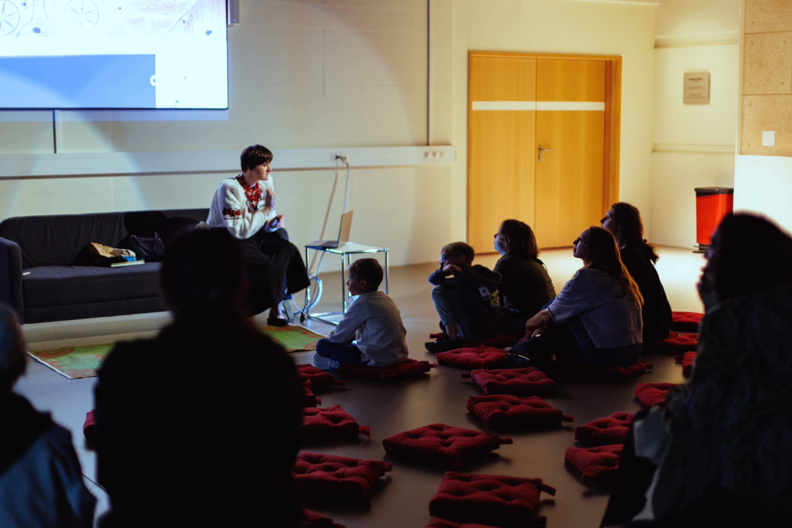 Children sitting on the floor in a room, listening to a speaker in front of a projection screen.