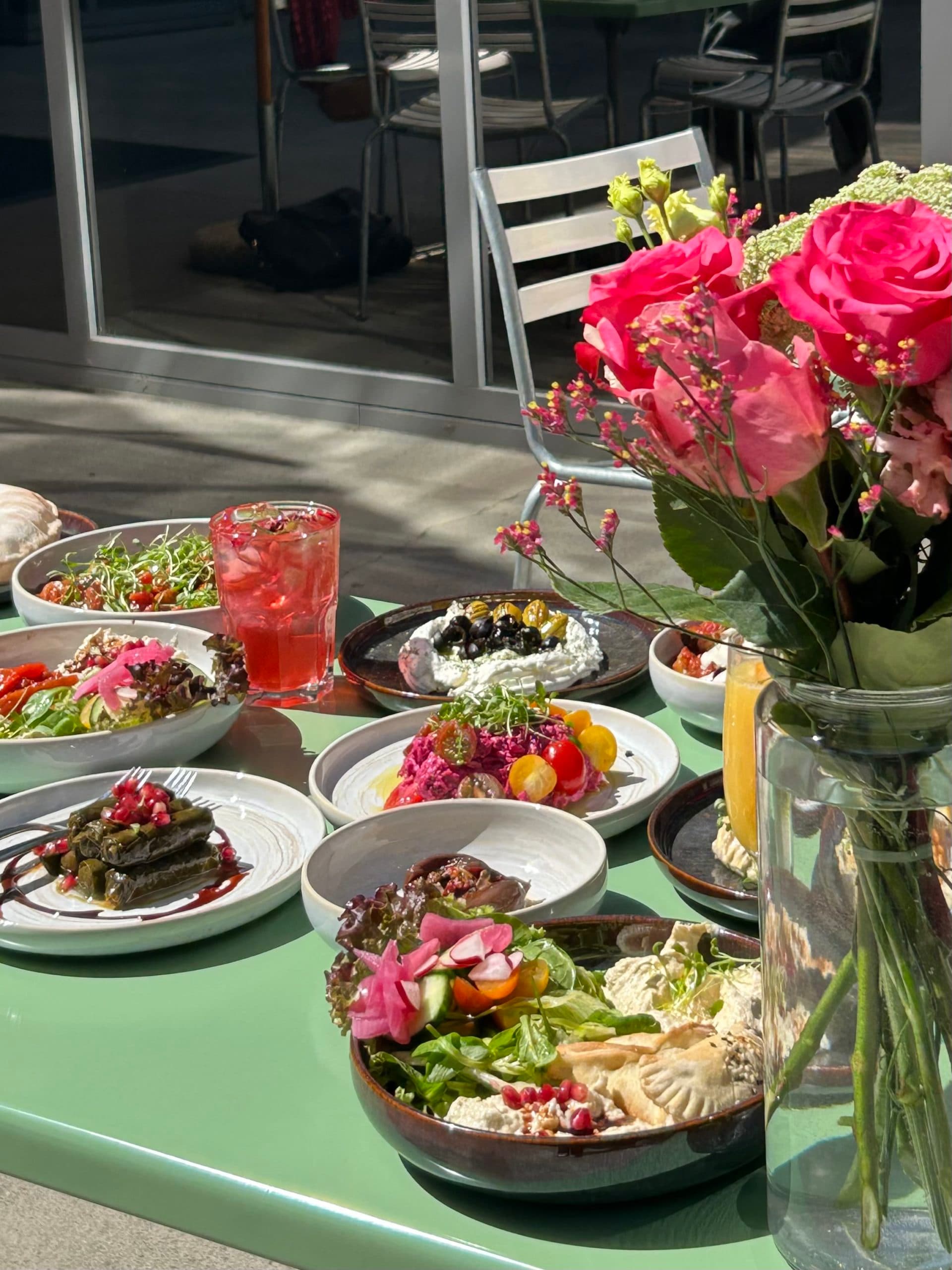 Green table outside on the sun with colored plated of food with a roses bouquet.