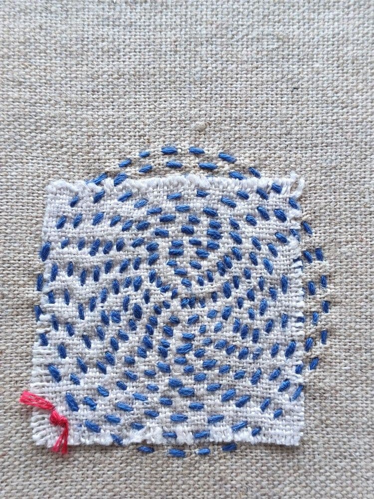 Stiche, die einen Kreis aus blauem Garn auf grauem Stoff bilden, der mit einem weißen Stoffquadrat geflickt ist.