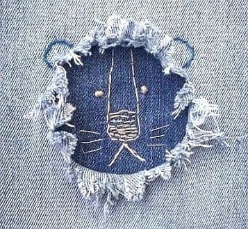 Nahaufnahme eines ausgefransten Lochs in einer hellen Jeans, durch das dunkelblauer Stoff mit einer weißen Zierstickerei zu sehen ist, die ein Tiergesicht in das Loch zeichnet.