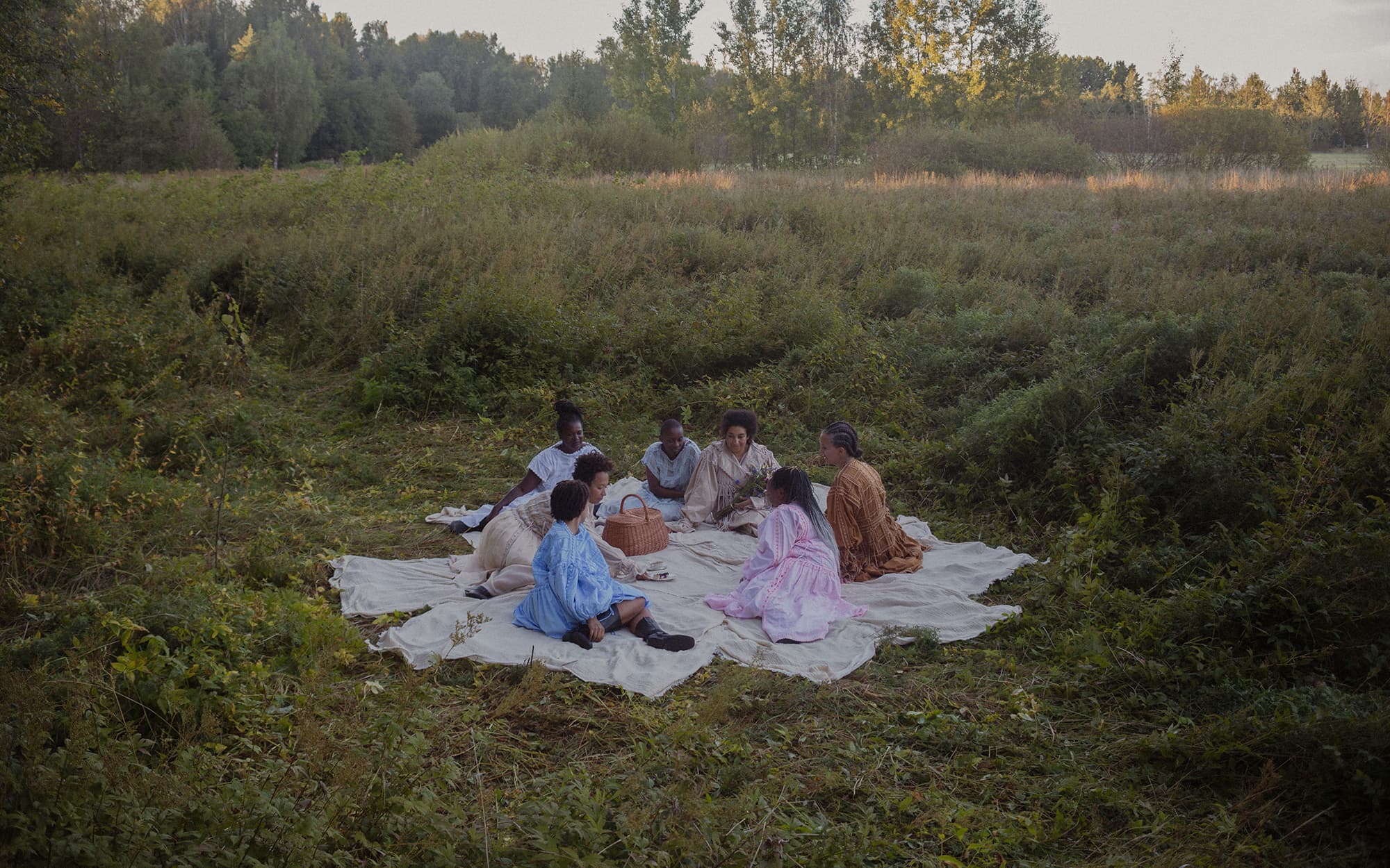 Women gathered together on some white tablecloths in the middle of a high-grass field for a picnic with a warm and calm atmosphere.