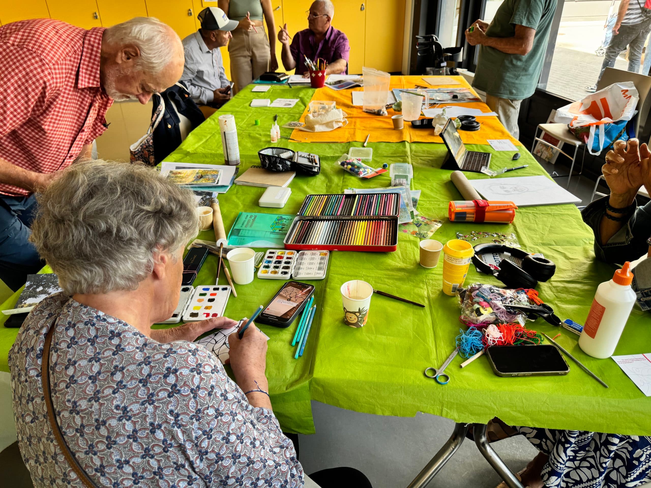 Des seniors sont assis autour d'une table avec une nappe verte durant un atelier de dessin. Des boîtes de crayons, des boîtes de peinture, des pinceaux et des fils de couleur sont disposés sur la table.