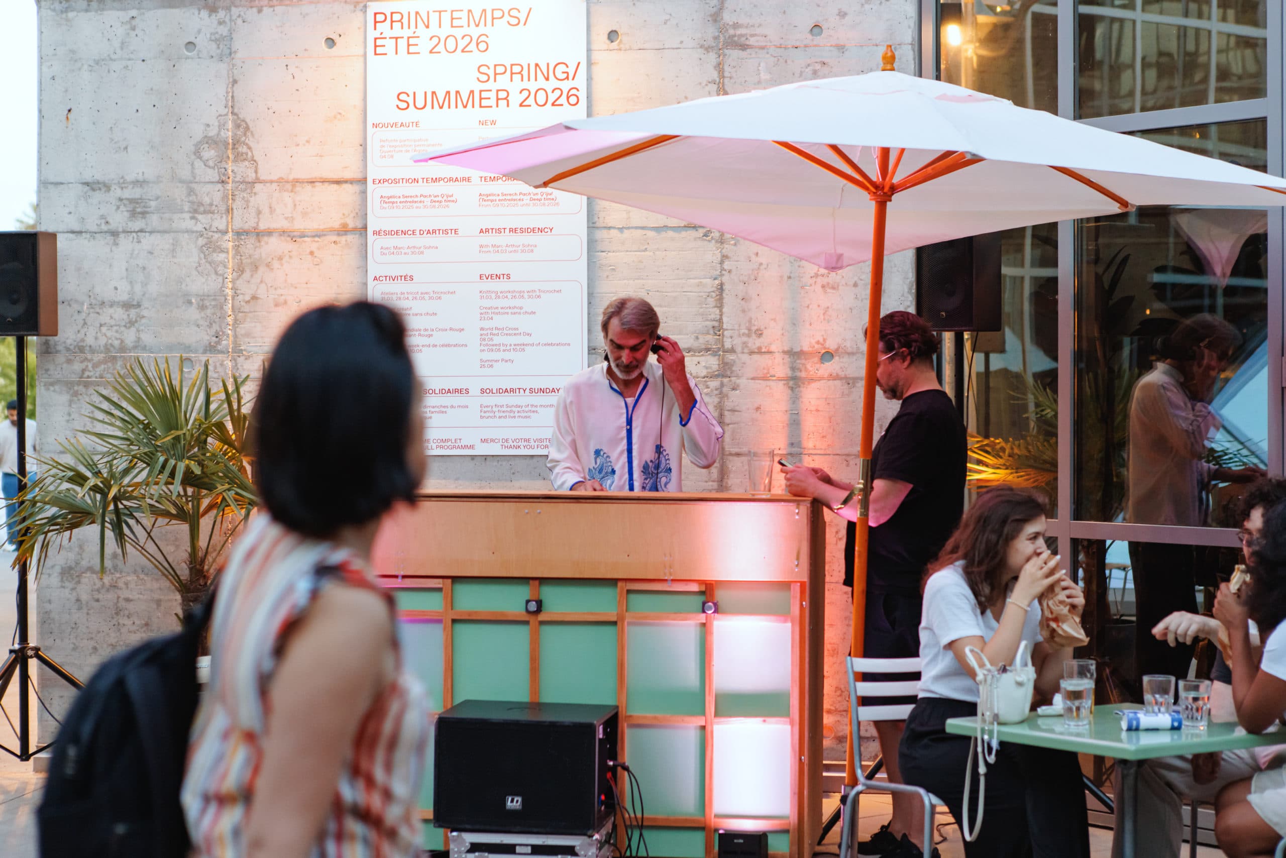 DJ behind a mixing desk at an outdoor terrace event under a large white umbrella, while guests chat and enjoy drinks at tables in a contemporary space lit by warm ambient lighting.