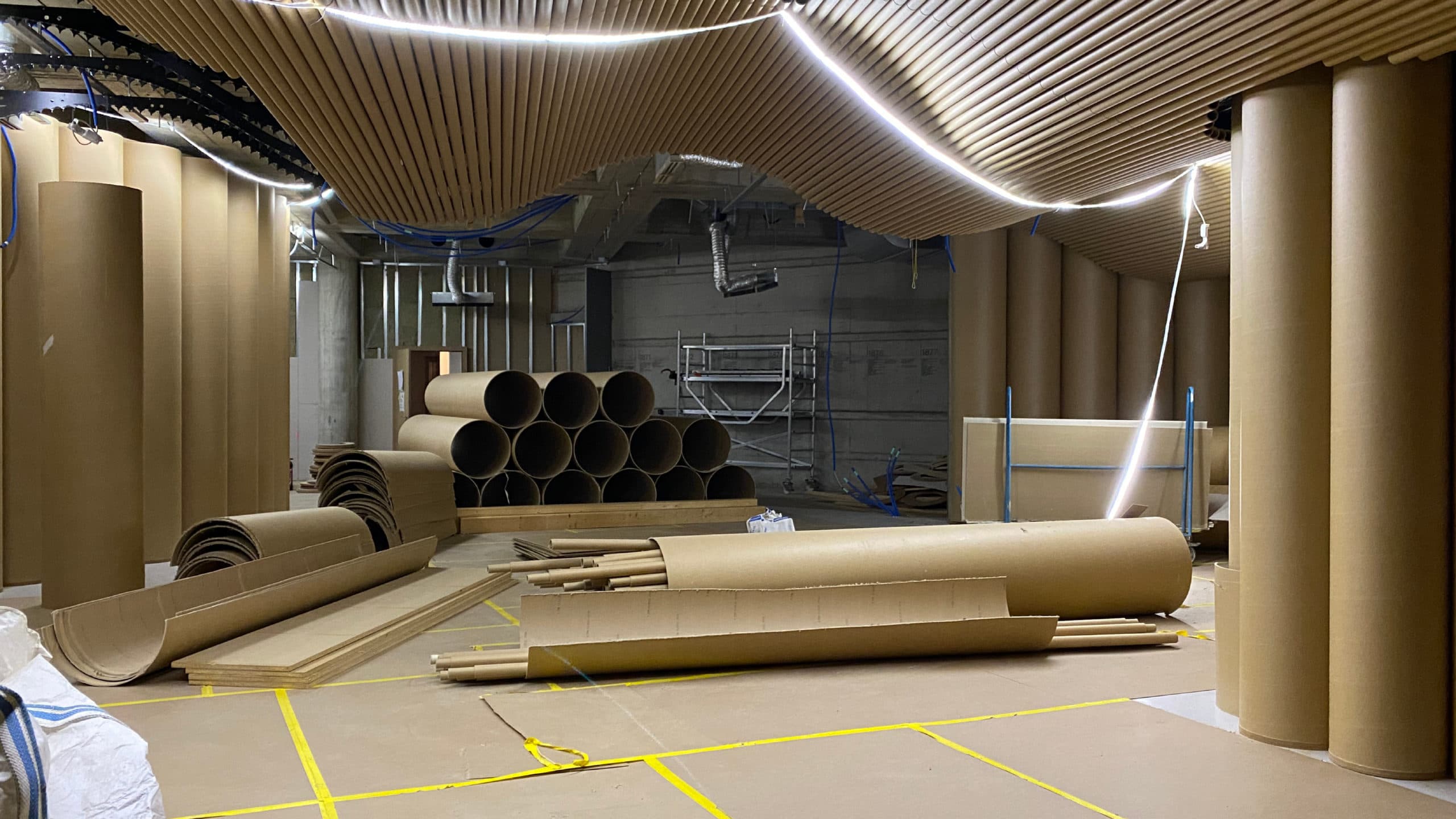 Une salle en travaux éclairée par une guirlande de leds puissante contenant de grands cylindres de cartons aux murs. Certains ont été démontés et sont empilés les uns sur les autres au sol.