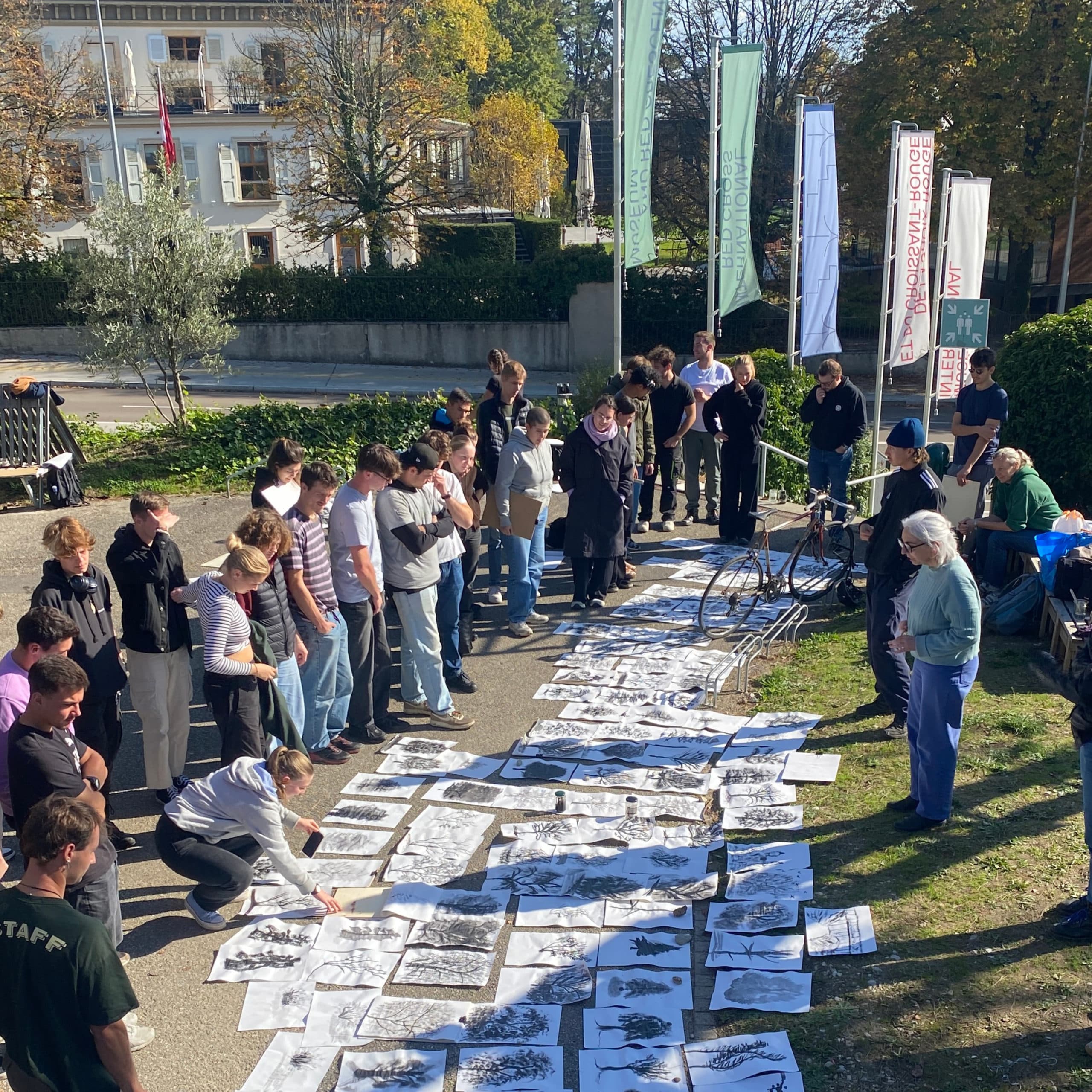Des participants rassemblés en extérieur examinent des plans, dessins et photographies étalés sur le sol pendant qu'une personne présente et commente les documents.